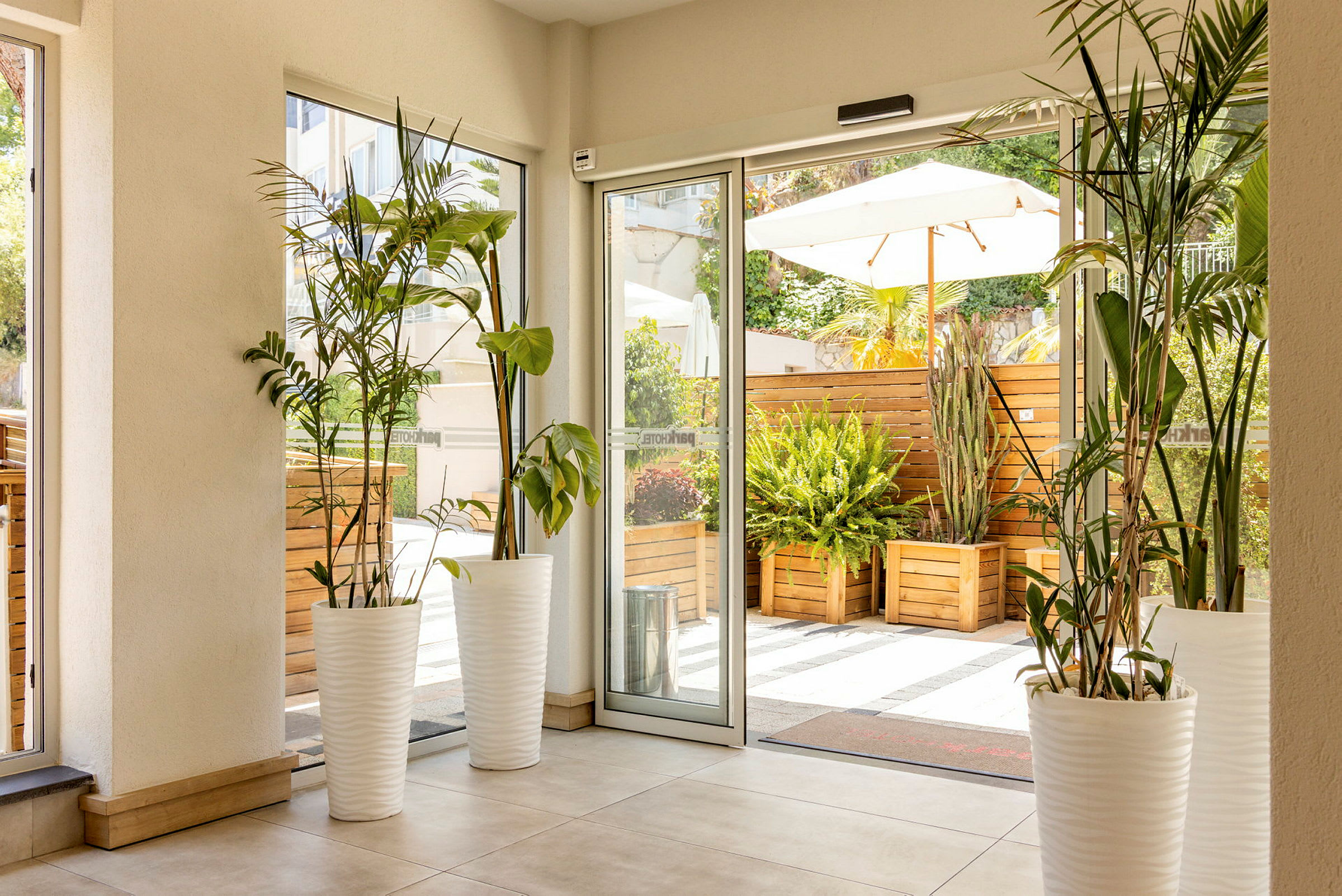 Interiør av et rom med planter i hvite potter ved siden av glassdører som åpner ut mot en terrasse med trekasser og frodig beplantning. (AI-generert tekst)