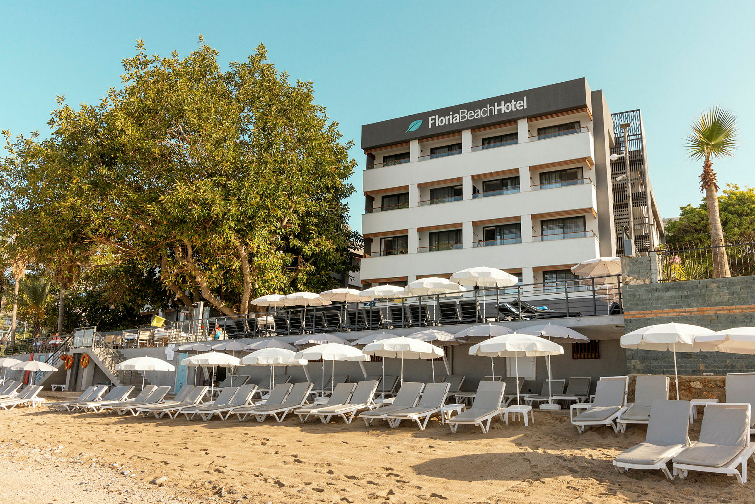 Hvit bygning med skiltet 'Floria Beach Hotel' og en sandstrand med solsenger og parasoller. (AI-generert tekst)