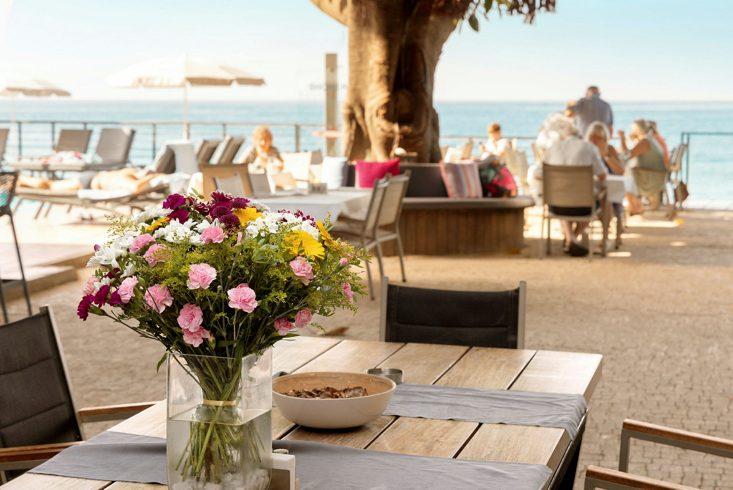 Et bord med en vase fylt med fargerike blomster, med folk som sitter ved andre bord i bakgrunnen nær havet. (AI-generert tekst)