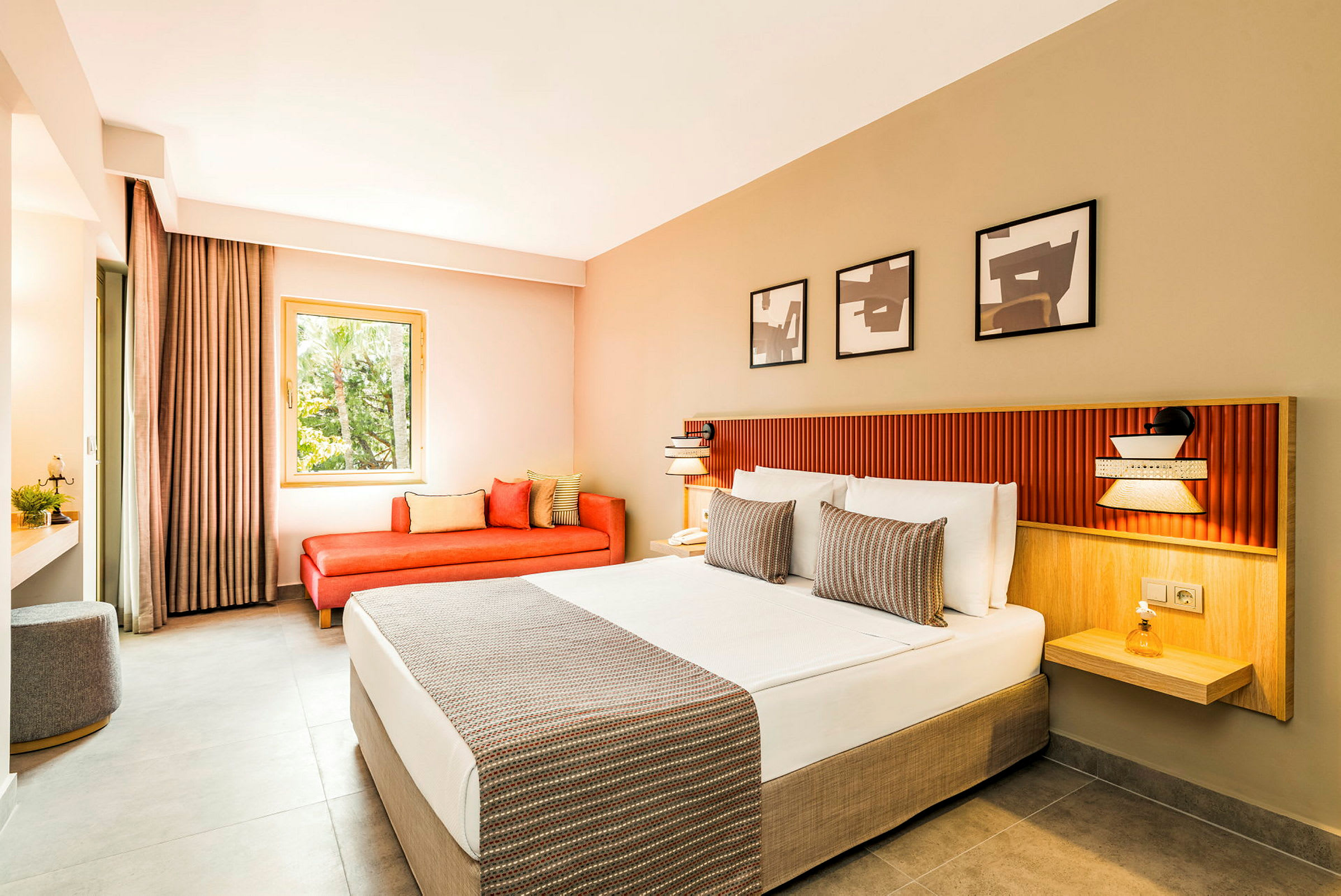Et moderne hotellrom med en bred seng med hvite laken og dekorative puter. Det er en rød sofa ved vinduet og tre kunstverk på veggen over sengen. (AI-generert tekst)