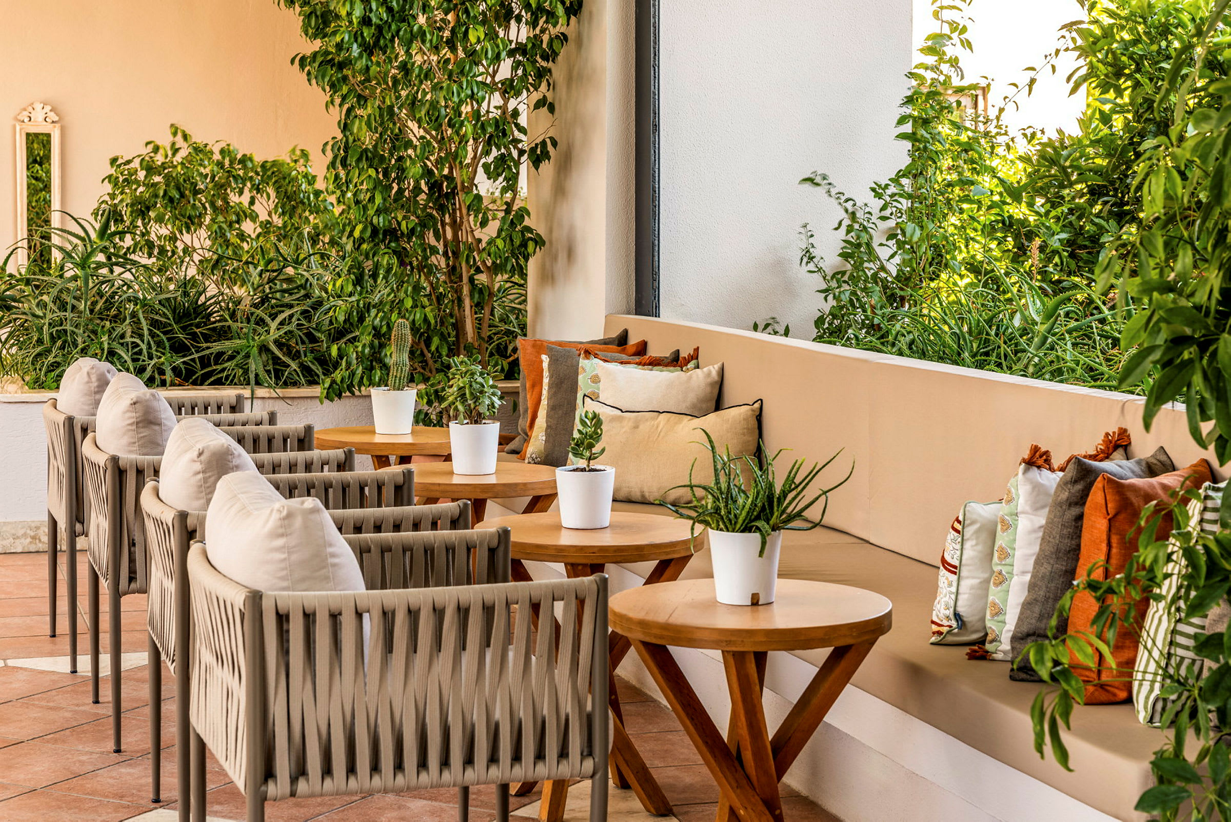 En terrasse med flere bord og stoler samt en sofa med fargerike puter. Planter og grønt omgir området. (AI-generert tekst)