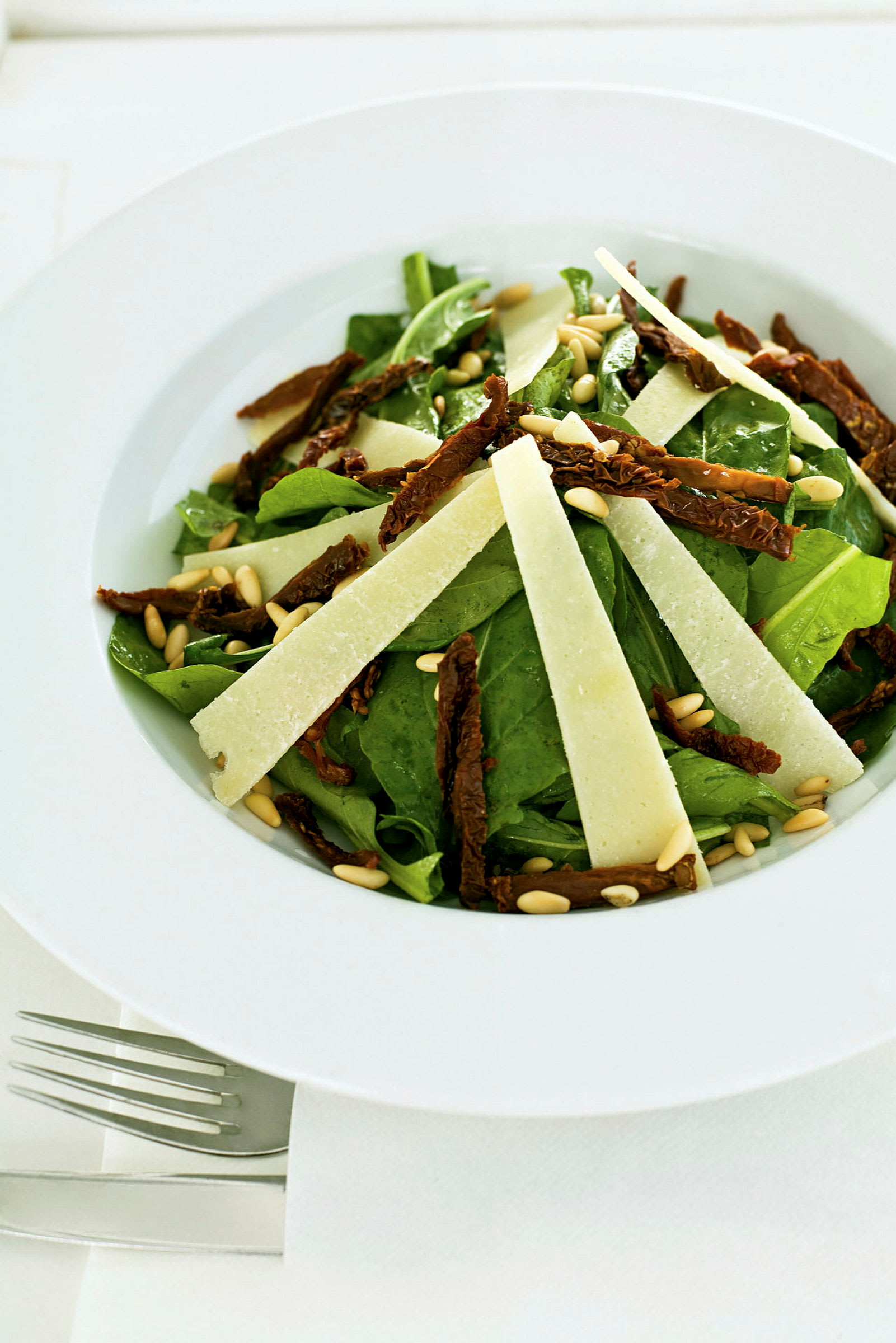 En tallerken med salat som inneholder grønne blader, soltørkede tomater, pinjekjerner og tynne osteskiver. (AI-generert tekst)