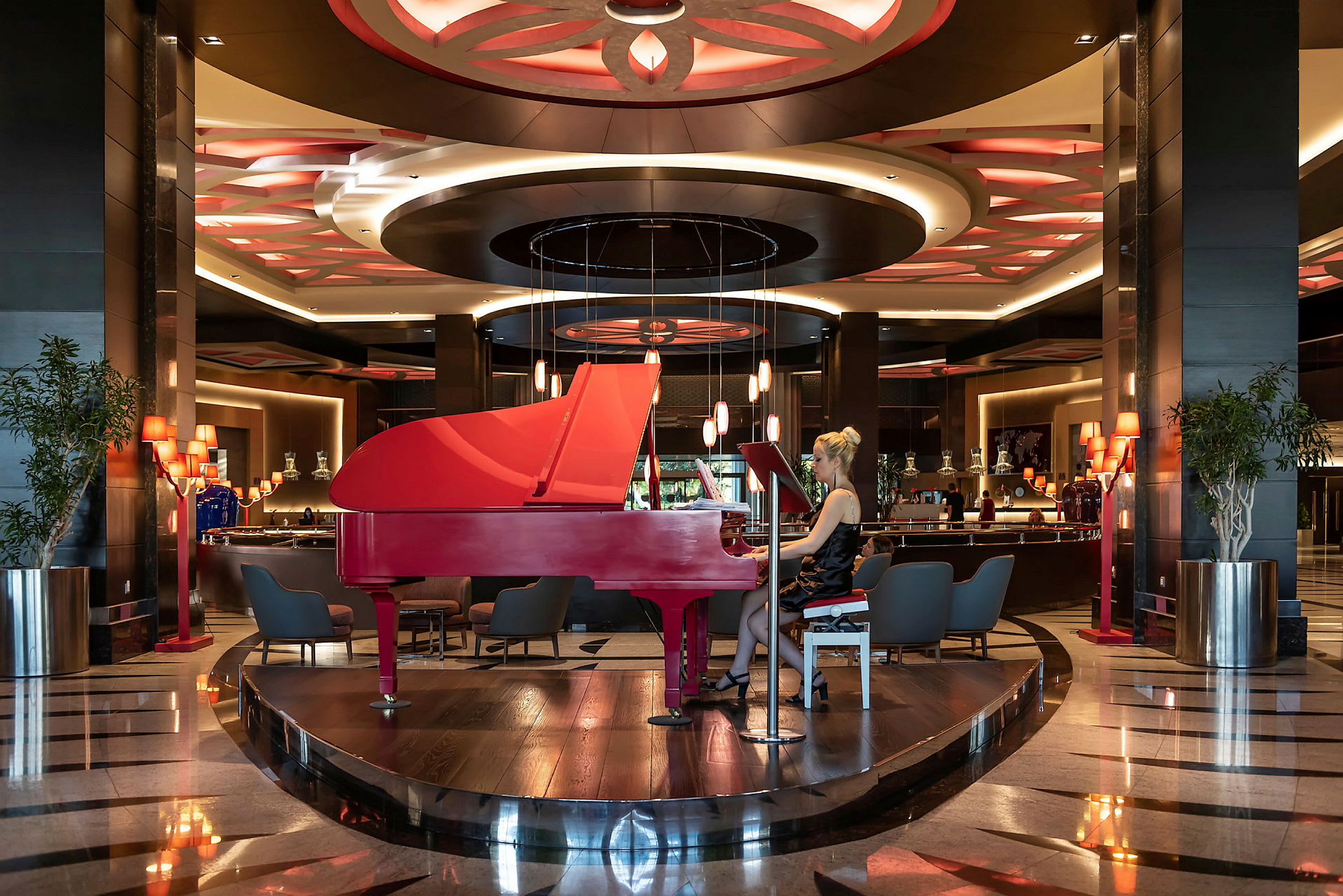 Et rødt flygel står sentralt plassert i en elegant hotellobby med dekorative taklamper. (AI-generert tekst)
