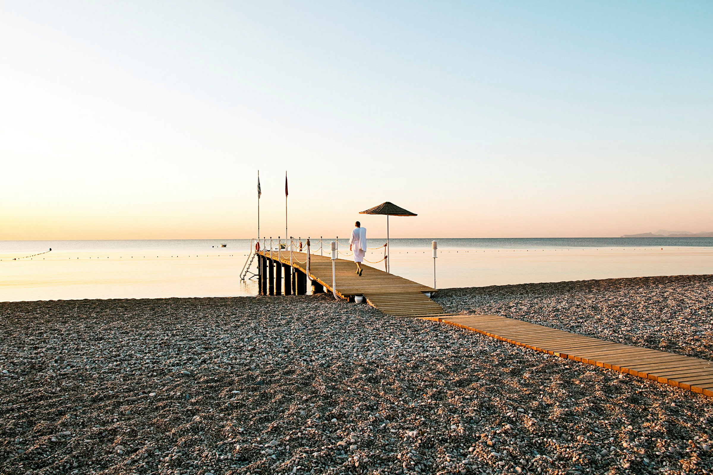 En steinstrand med en lang trebrygge som strekker seg ut i et rolig hav ved solnedgang. (AI-generert tekst)