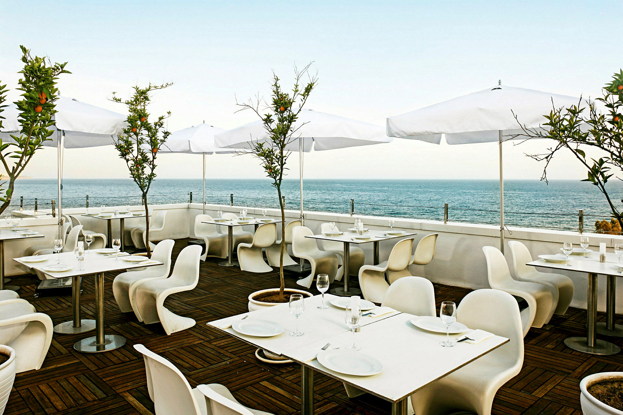 Restaurant med uteservering, hvite bord og stoler samt hvite parasoller. Bakgrunn med utsikt over havet. (AI-generert tekst)