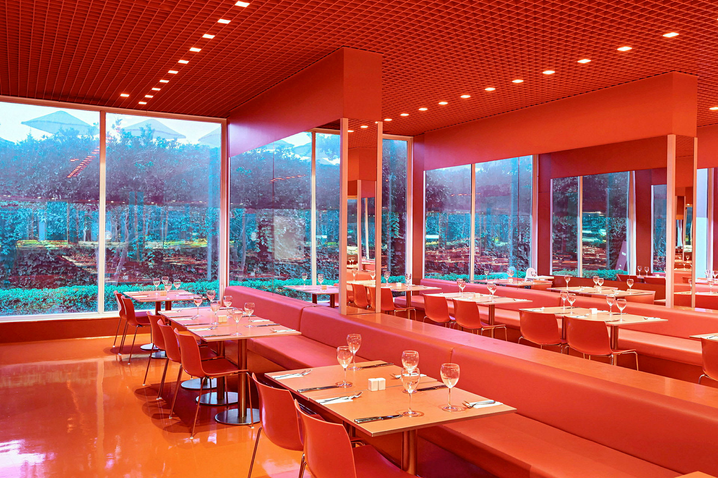 En moderne restaurant med røde møbler og store glassvinduer som vender mot en frodig hage. (AI-generert tekst)