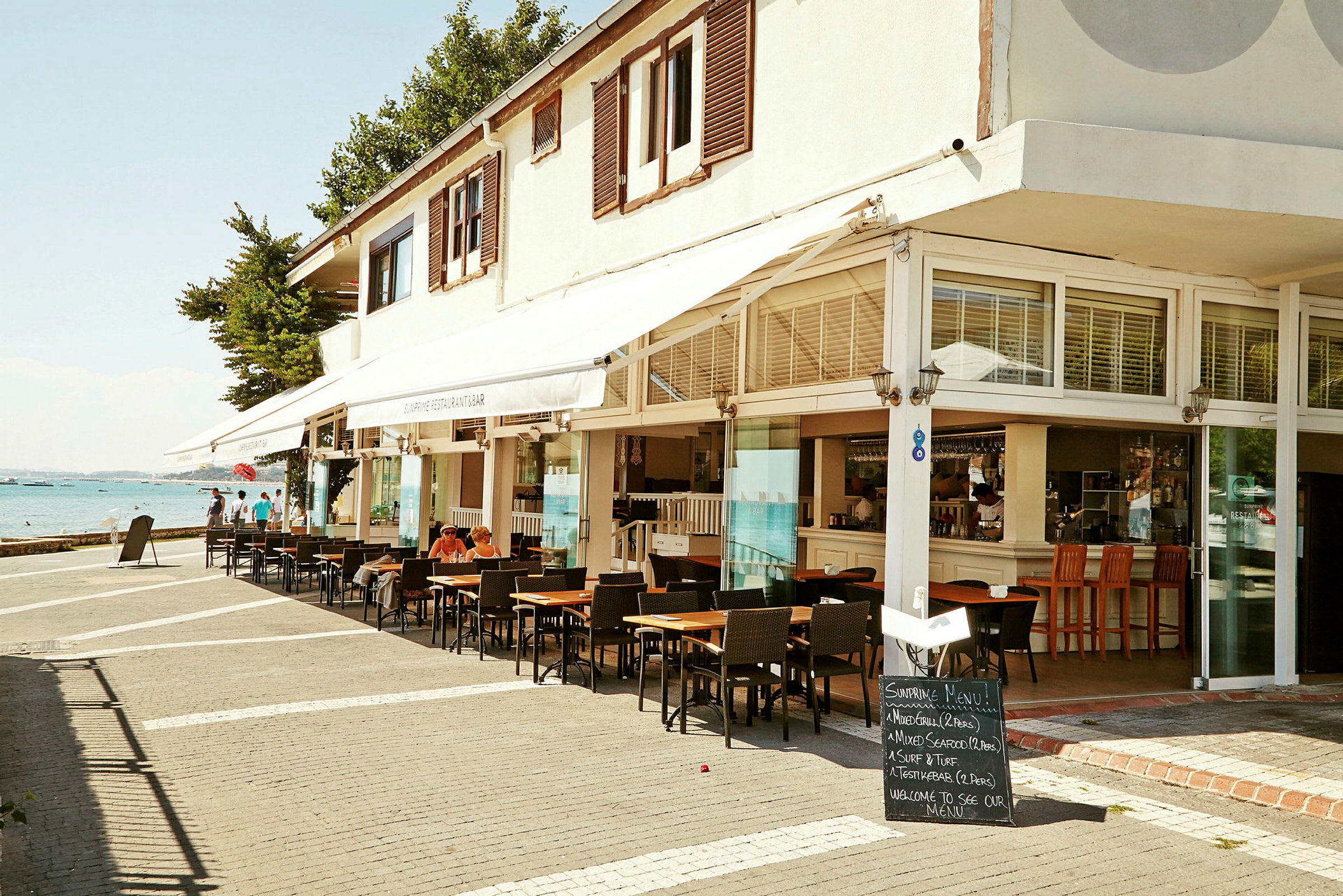 En kafé eller restaurant med sitteplasser utendørs og utsikt mot havet. (AI-generert tekst)