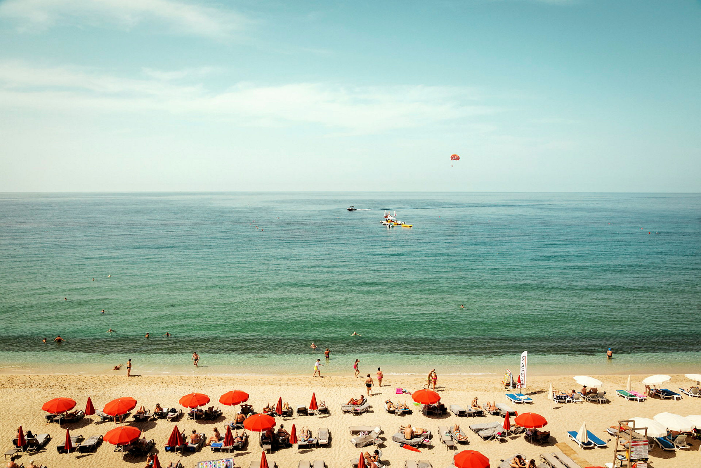 En strandscene med røde parasoller, solsenger og mennesker ved vannet. (AI-generert tekst)