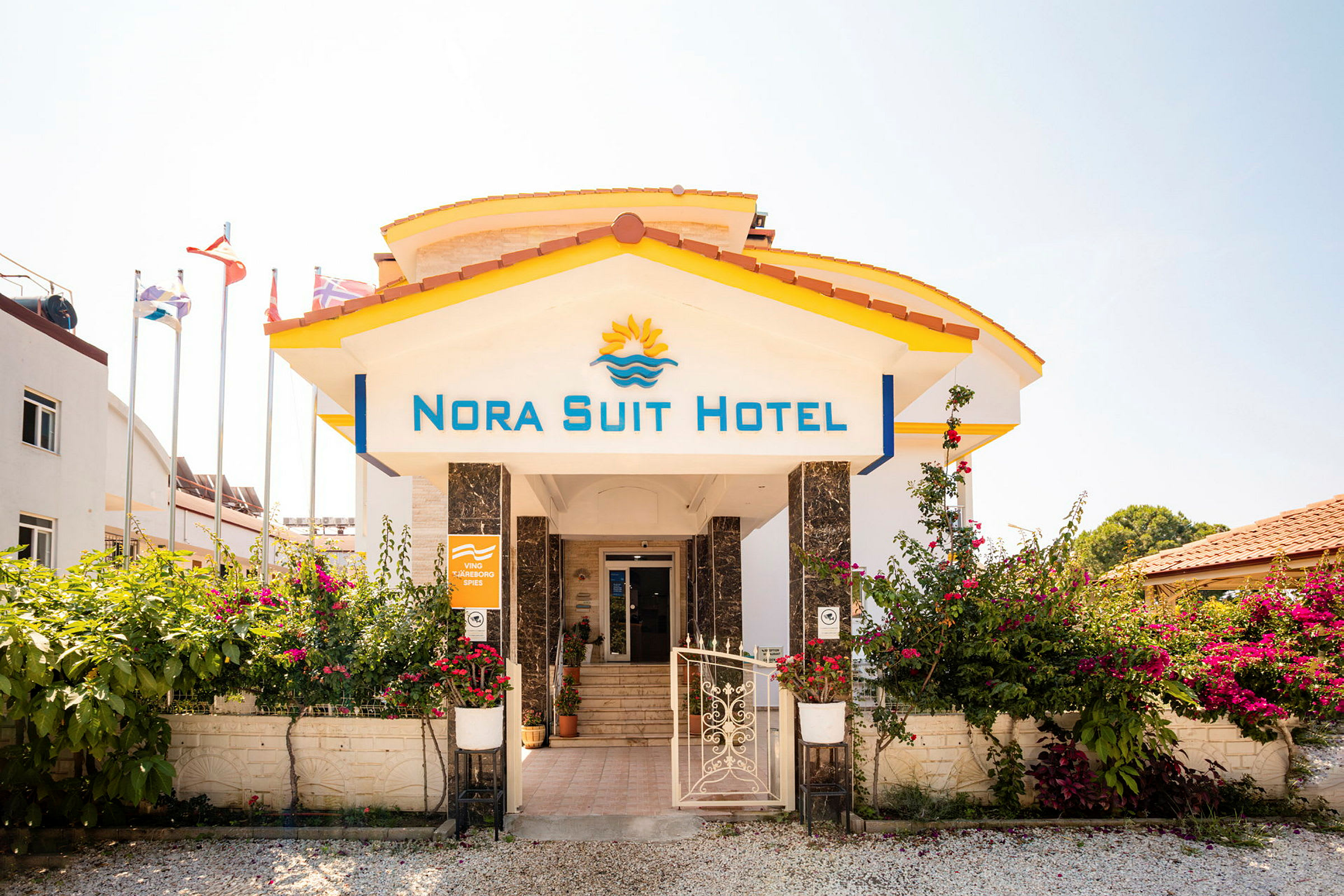 Inngangen til Nora Suit Hotel med blomster og flagg i bakgrunnen. (AI-generert tekst)