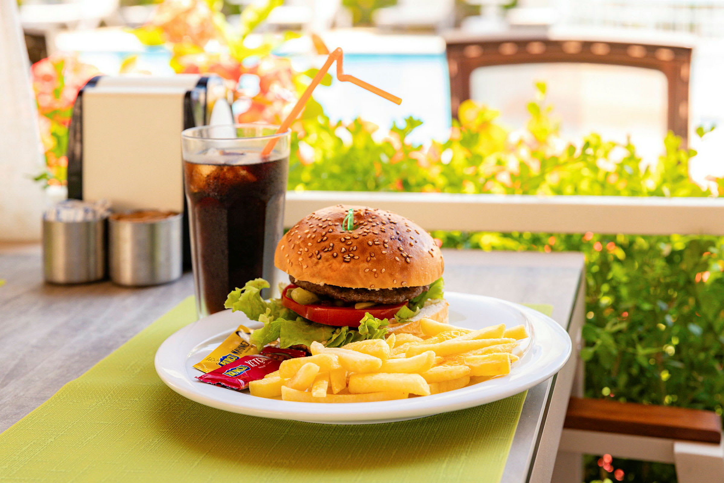 En tallerken med en hamburger, pommes frites og krydderposer, samt et glass brus, står på et bord utendørs. (AI-generert tekst)