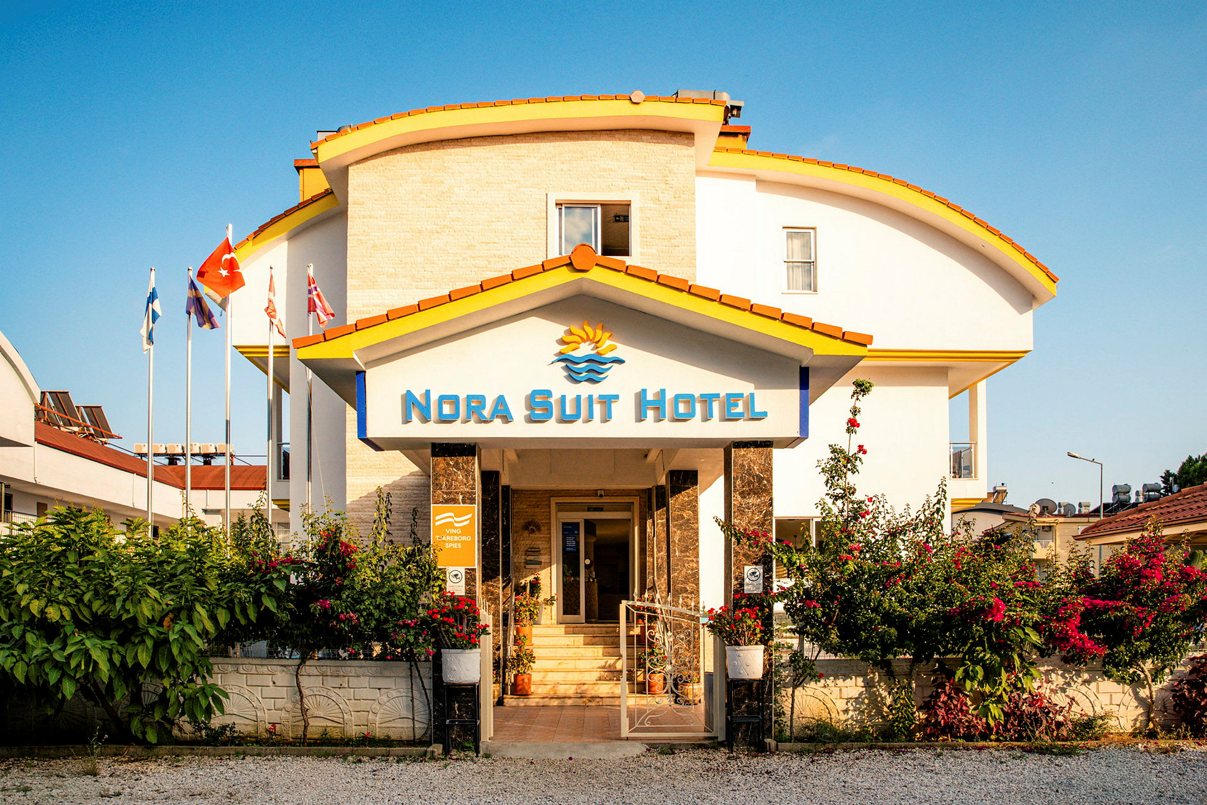 Bygningen til Nora Suit Hotel med en inngang omgitt av blomster og flagg. (AI-generert tekst)