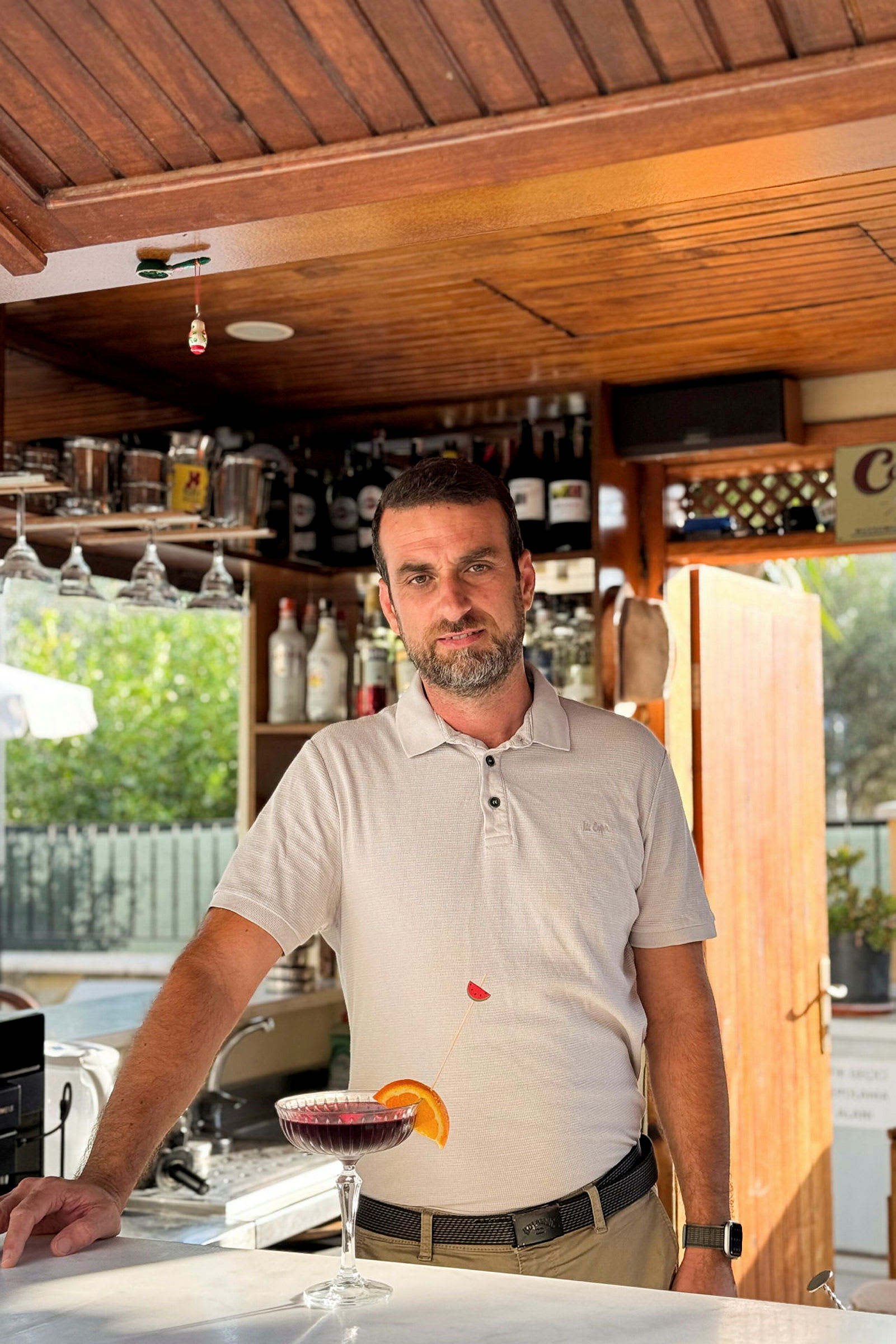 En hvit skjorte ved en bar med hyller fylt med flasker i bakgrunnen, samt et cocktailglass med en farget drikk foran seg. (AI-generert tekst)