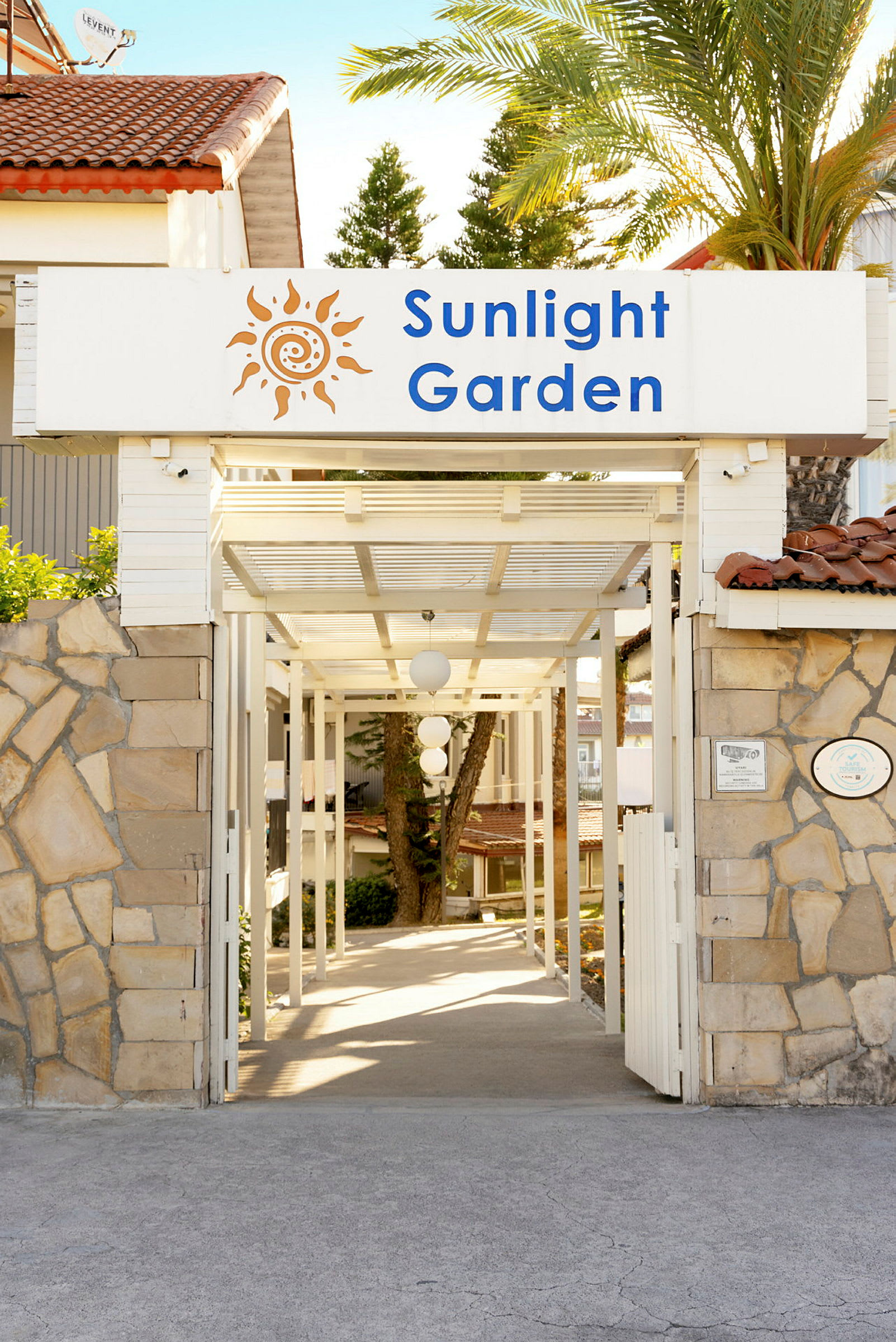 Inngang med skilt som sier 'Sunlight Garden', omgitt av steinmur og tropiske trær. (AI-generert tekst)
