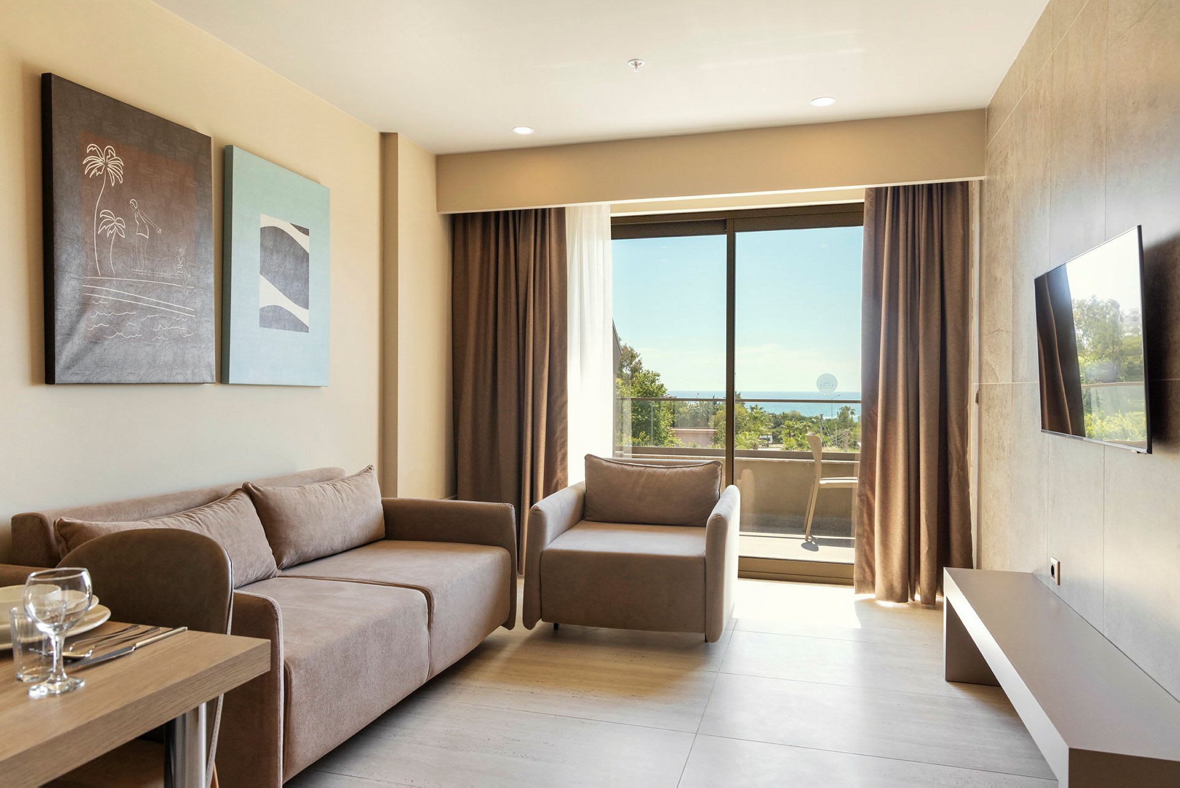 Et lyst hotellrom med en sofa, lenestol, malerier på veggen, et bord med glass og tallerkener, samt en balkong med utsikt mot havet gjennom store glassvinduer. (AI-generert tekst)