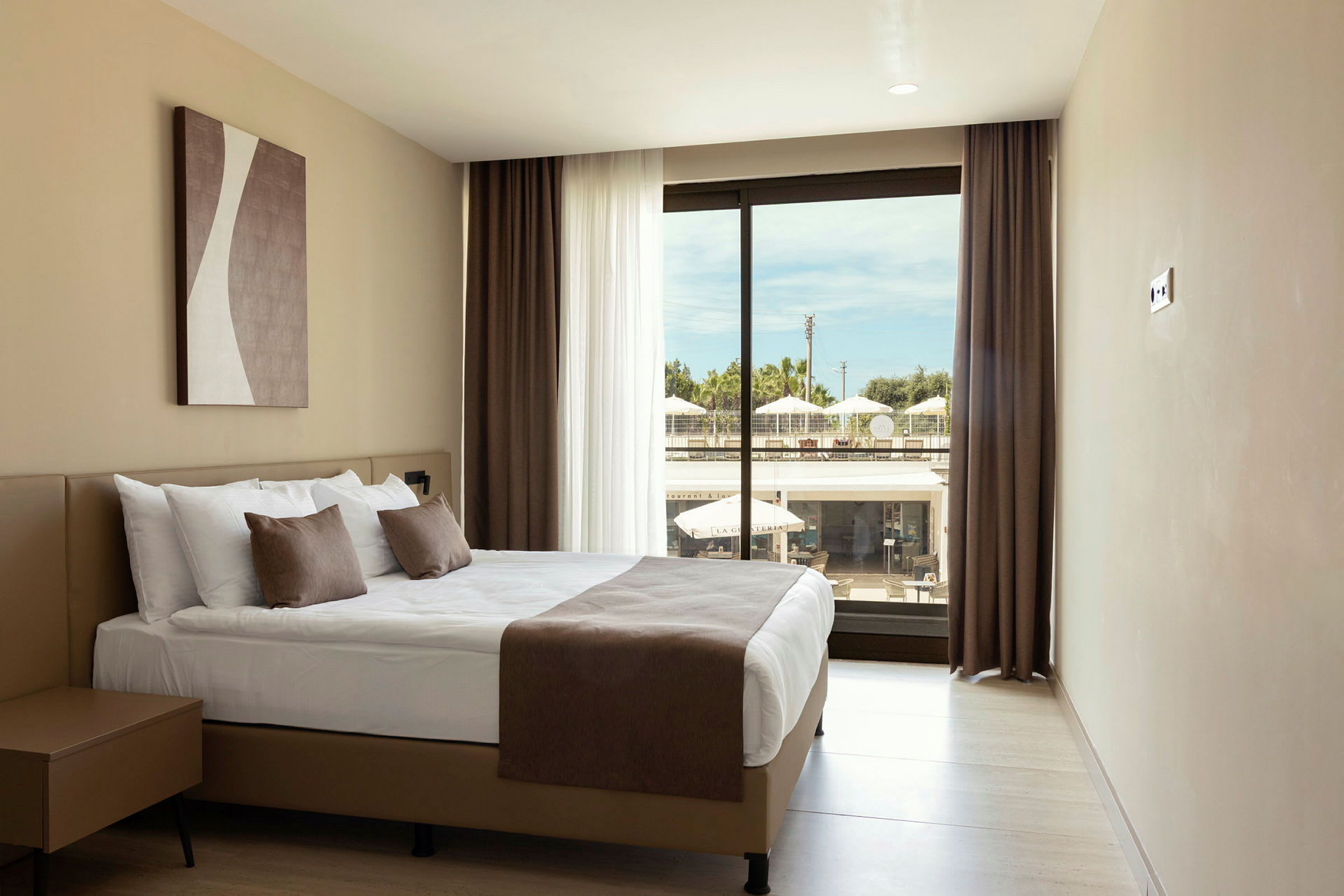 Et hotellrom med en dobbeltseng med hvitt sengetøy og brune dekorative puter. Et stort vindu med utsikt mot en terrasse utenfor. (AI-generert tekst)