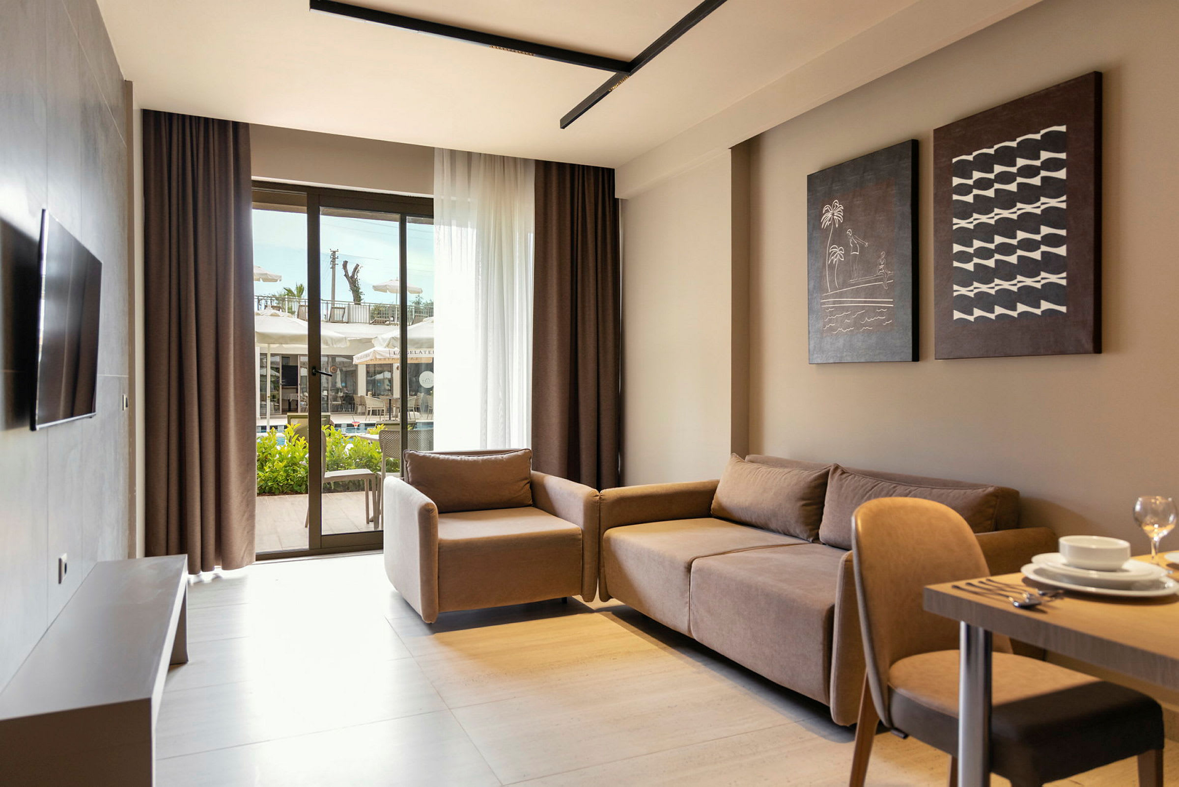 Et moderne hotellrom med sofa, lenestol og bord sett fra innsiden. Store glassdører i bakgrunnen viser utsikten mot en terrasse og svømmebasseng. (AI-generert tekst)