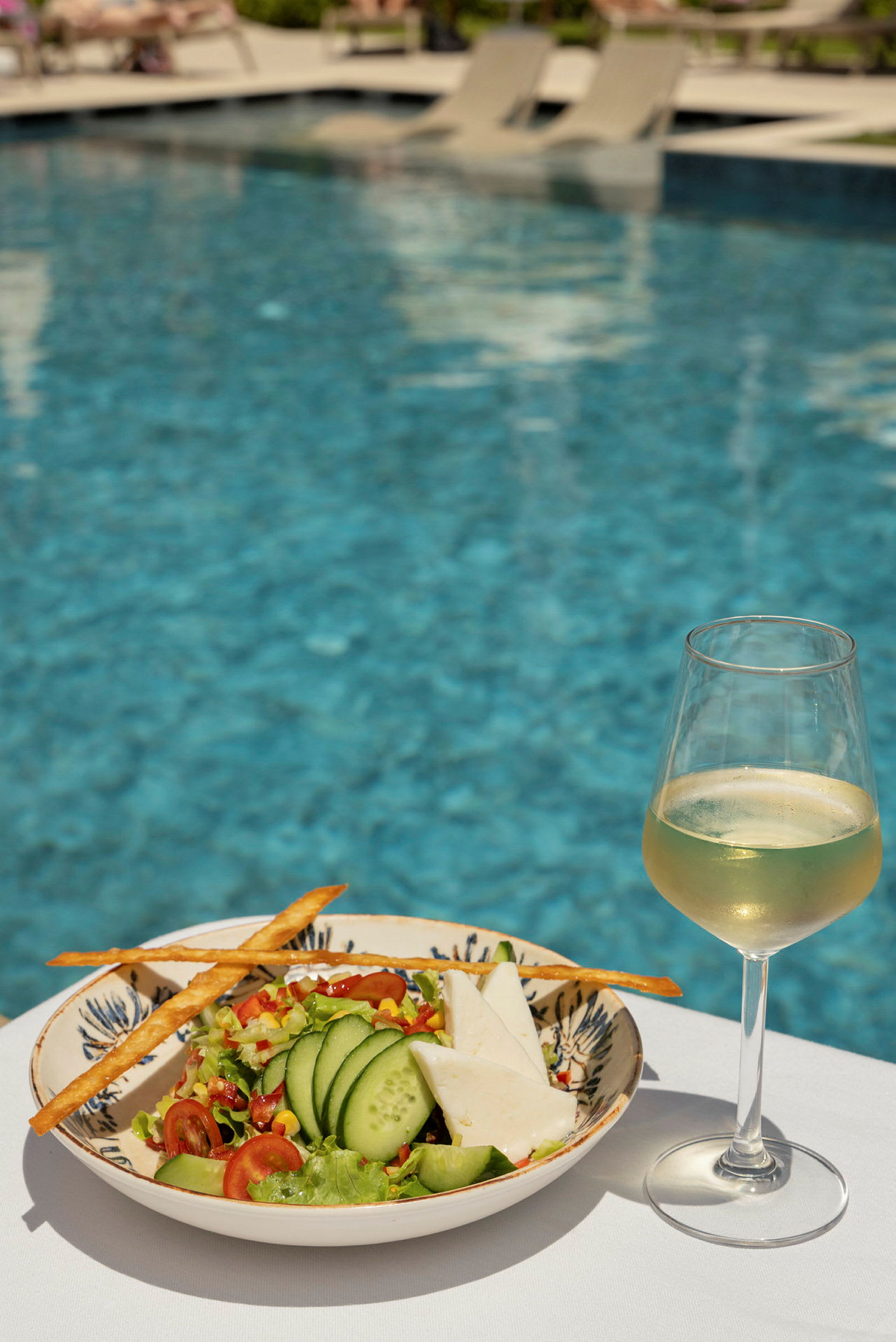 En salat med agurk og ost samt et glass hvitvin står på et bord med et basseng i bakgrunnen. (AI-generert tekst)