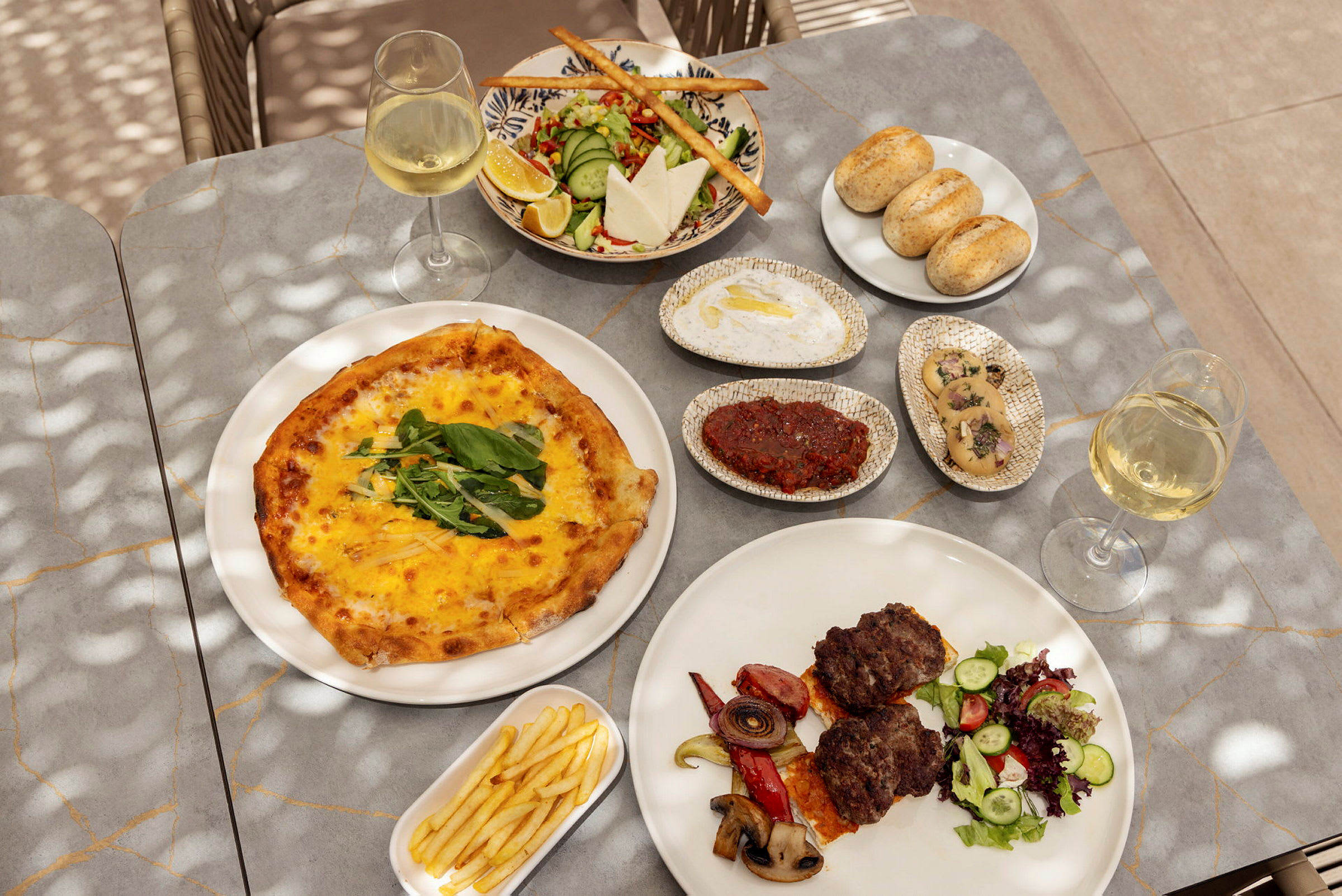 Et bord med forskjellige retter, inkludert pizza, salat, brød, kjøtt med grønnsaker, pommes frites og glass med hvitvin. (AI-generert tekst)