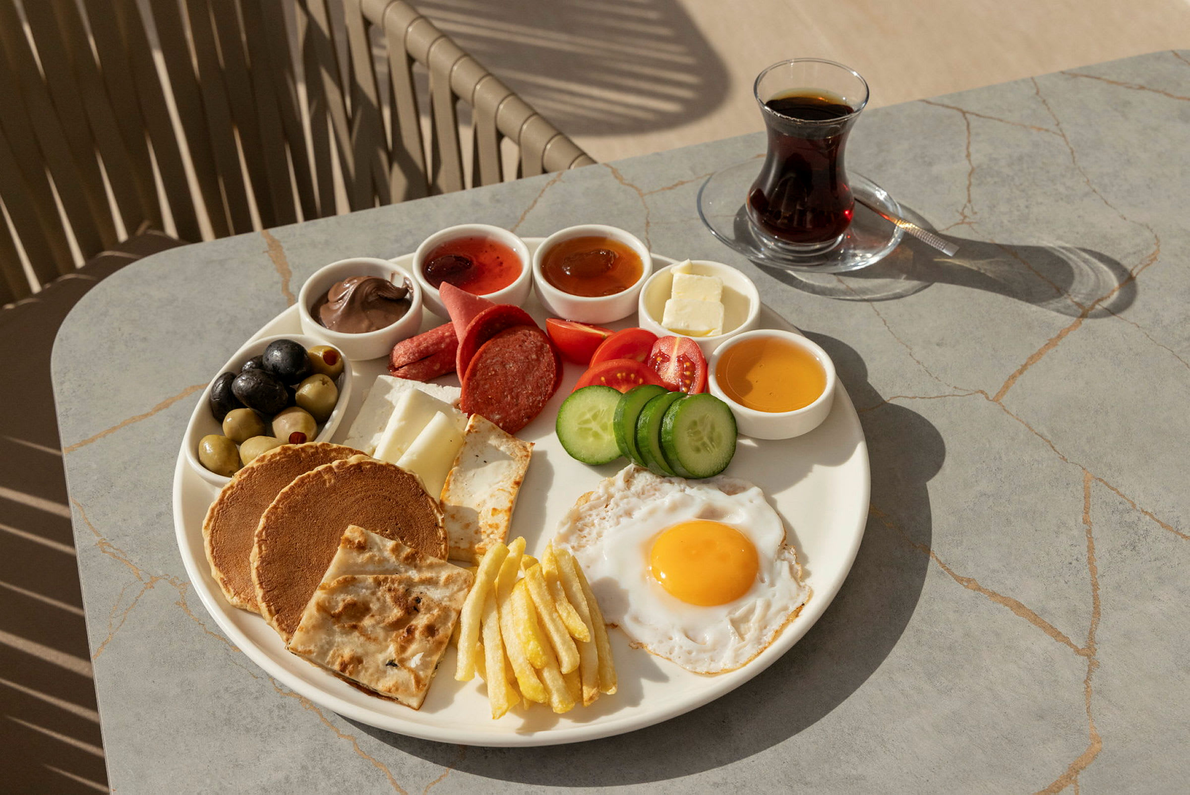 En tallerken med frokostmat som inneholder blant annet et stekt egg, grønnsaker, brød og små skåler med forskjellige tilbehør. Ved siden av står et glass te. (AI-generert tekst)