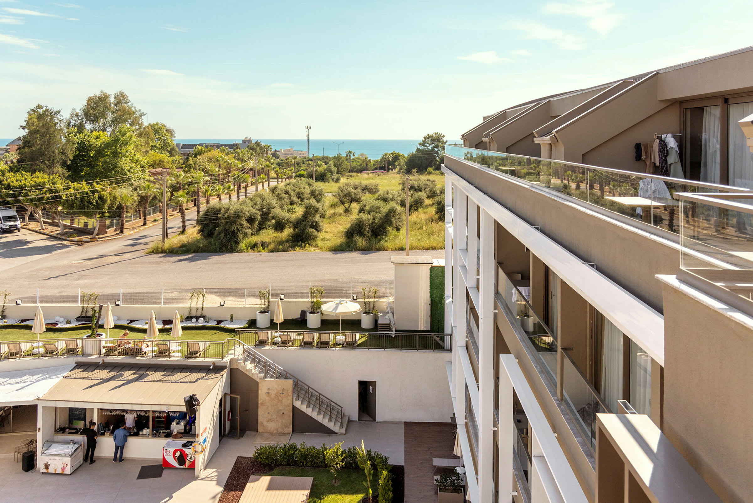 Et hotell med moderne arkitektur og balkonger, ved siden av et trebevokst område og en vei. I bakgrunnen kan havet sees. (AI-generert tekst)