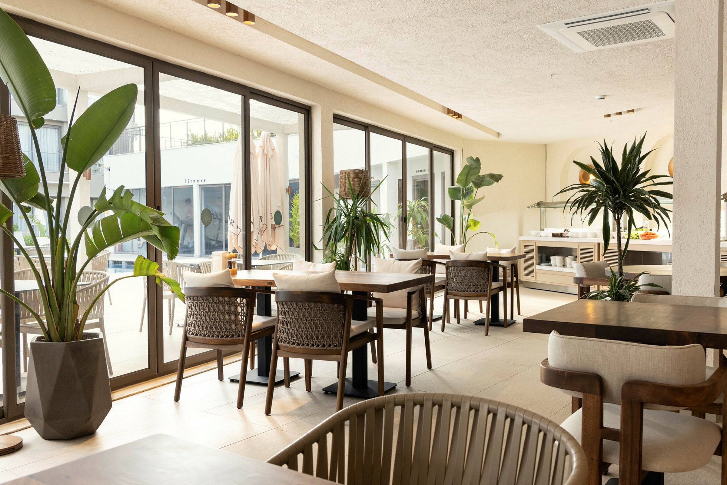 Restaurantområde med bord og stoler, omgitt av store vinduer og grønne planter. (AI-generert tekst)