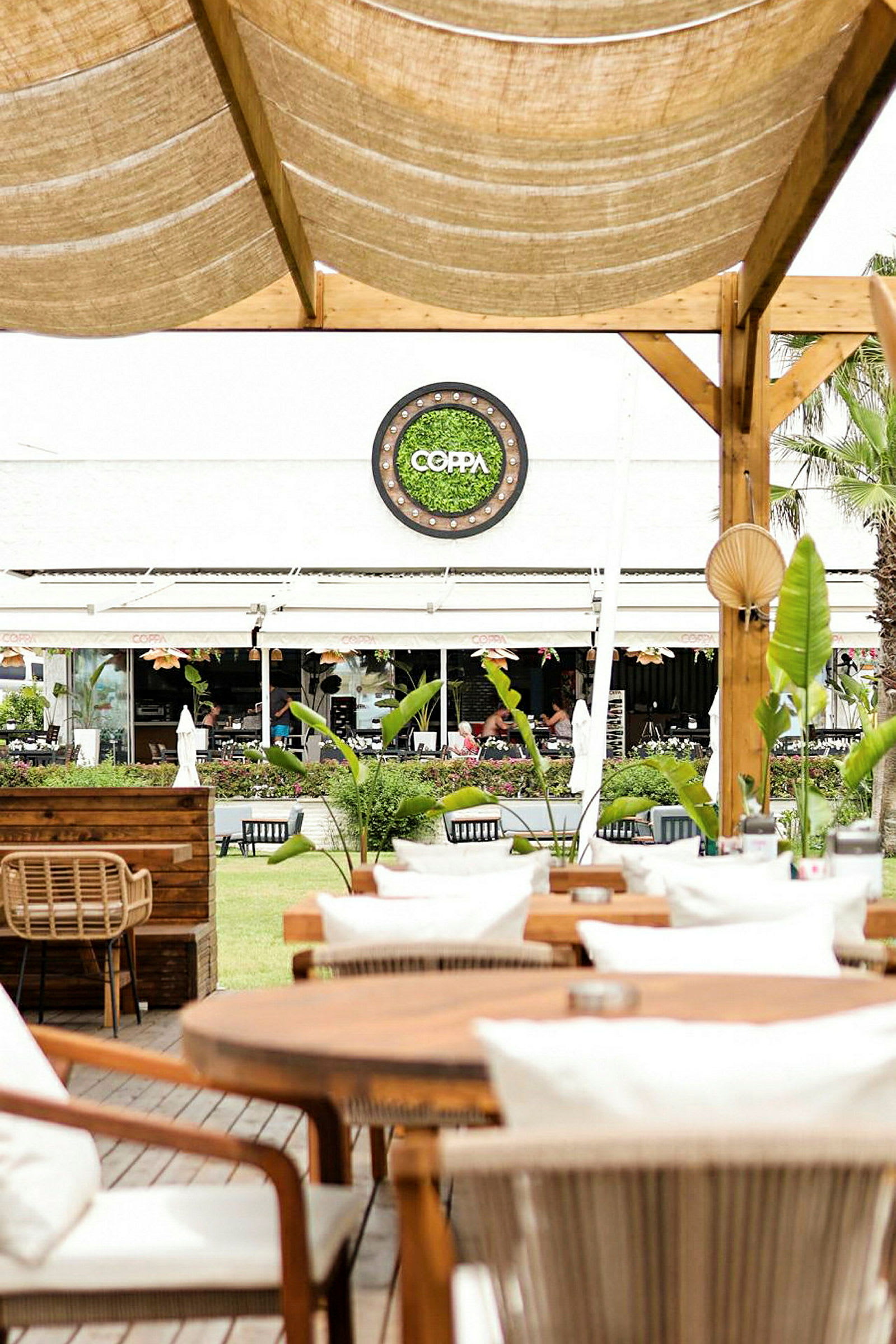 Utendørs servering med trebord og stoler, hage og restauranten Coppa i bakgrunnen. (AI-generert tekst)