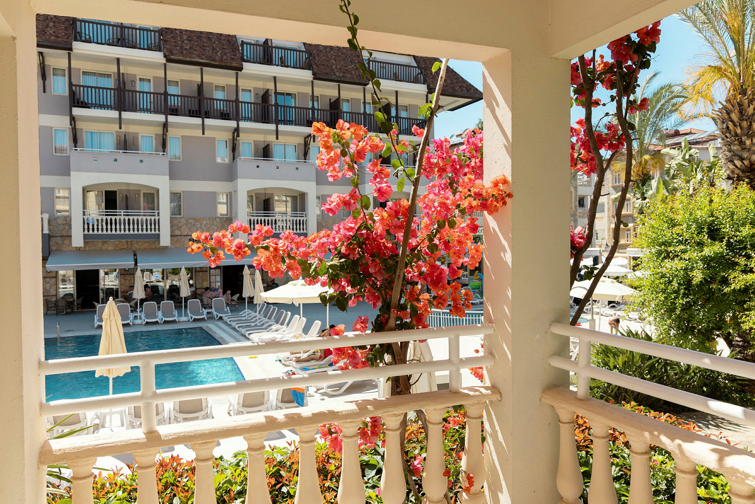 Utsikt fra en terrasse mot et basseng omgitt av solsenger, med blomstrende bougainvillea i forgrunnen. (AI-generert tekst)