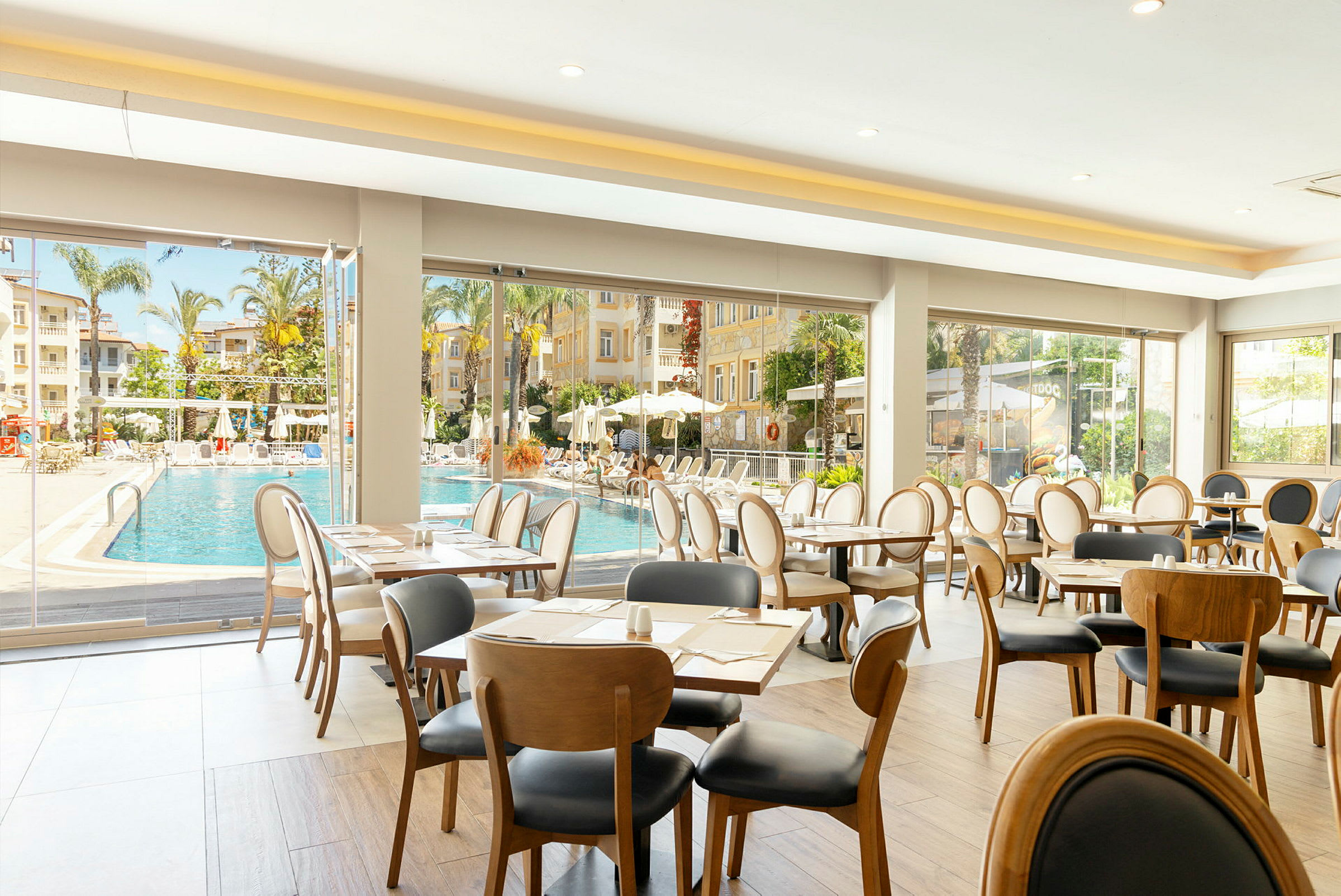 Interiør av restaurant med utsikt mot bassengområde og palmer. (AI-generert tekst)