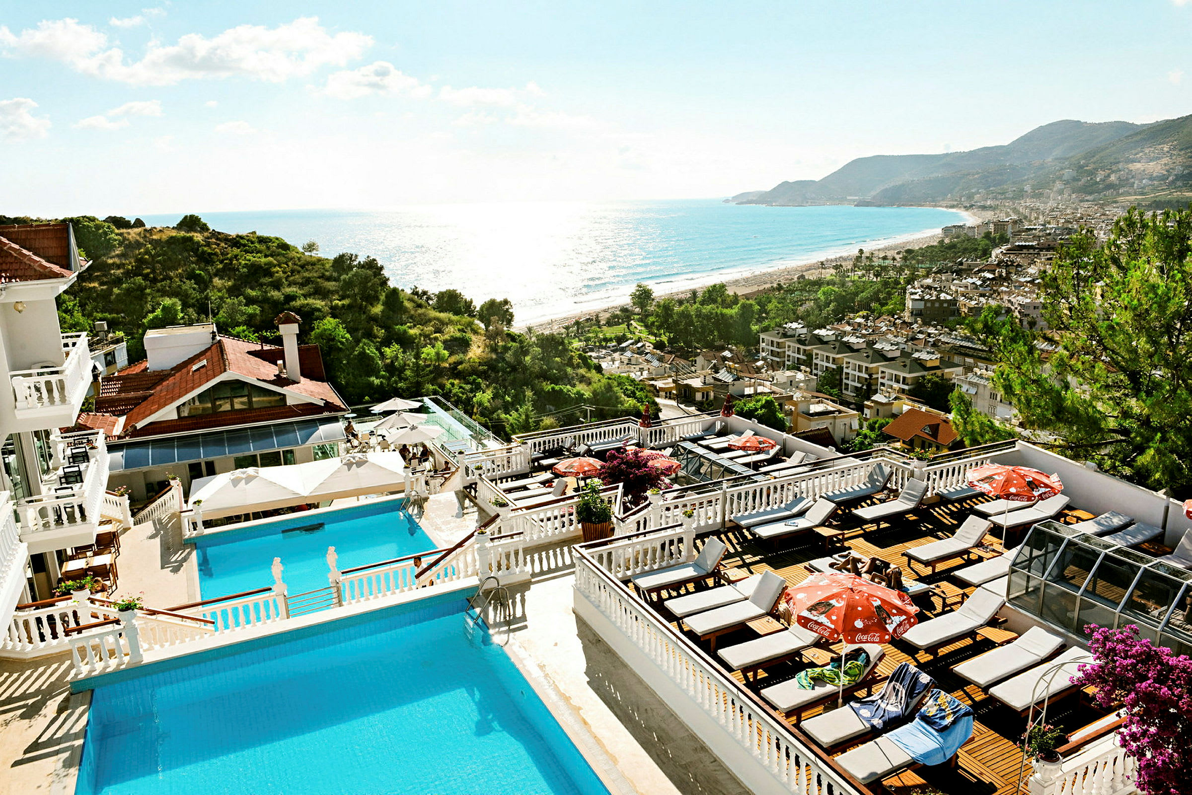 Bilde av et hotellområde med bassenger og solterrasser, omgitt av grøntområder og utsikt over havet og en strand. (AI-generert tekst)