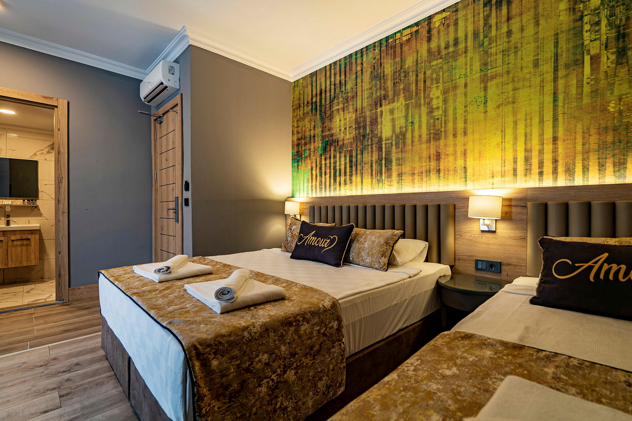Et hotellrom med to senger, luftig dekorert med gylne aksenter og en vegg med moderne tekstur bak sengene. (AI-generert tekst)