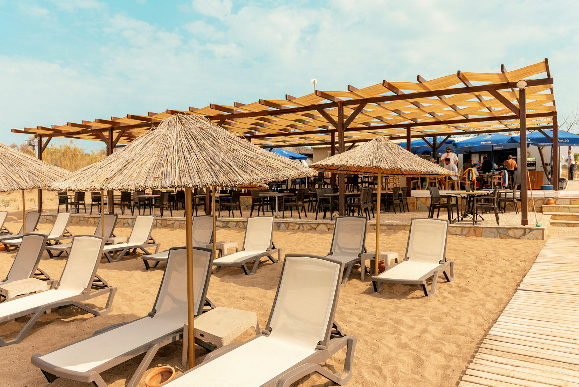 Strand med solsenger og stråparasoller foran en restaurant med åpen tak. (AI-generert tekst)