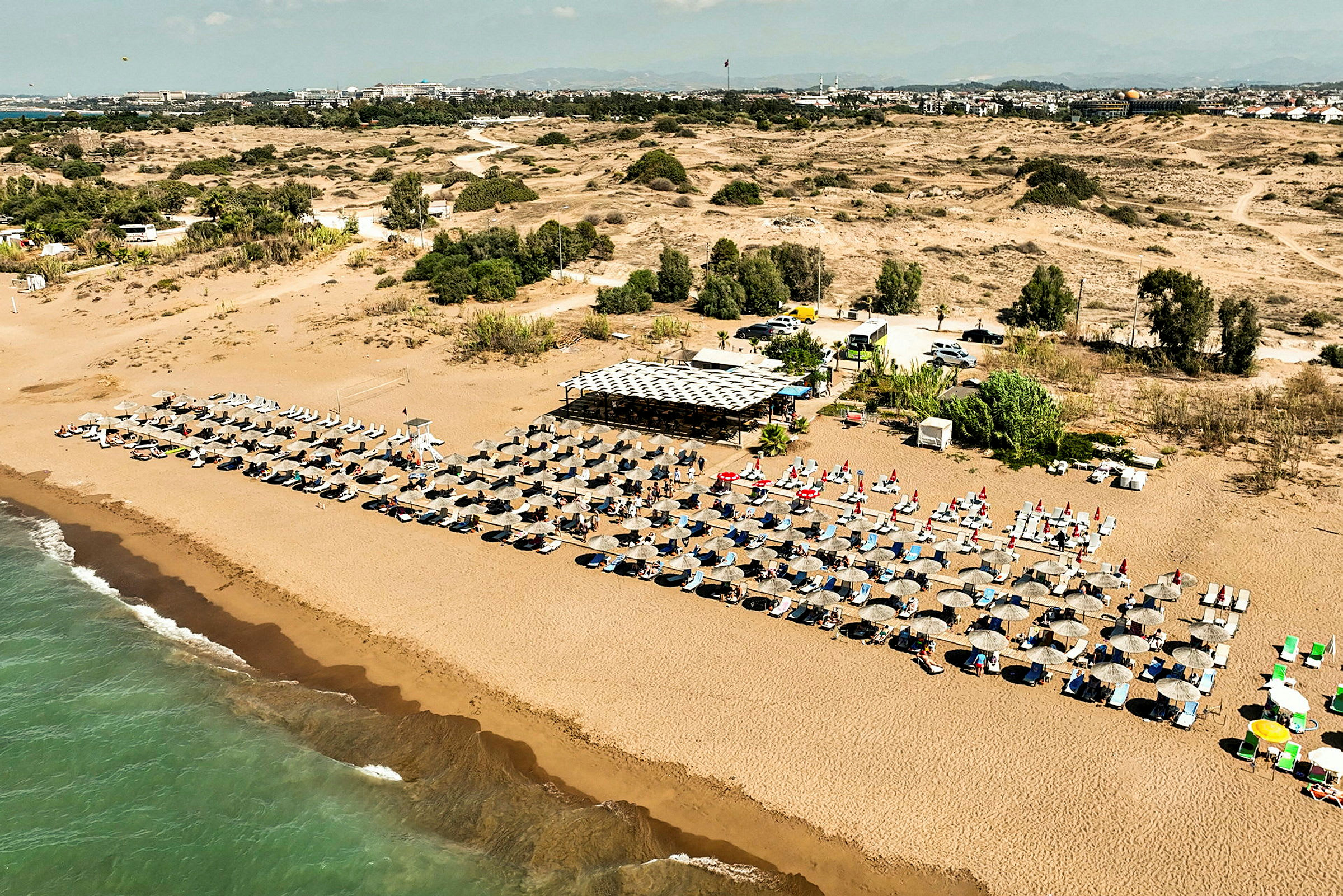 En lang sandstrand omgitt av rader med solsenger og parasoller med en strandkafé i bakgrunnen. (AI-generert tekst)