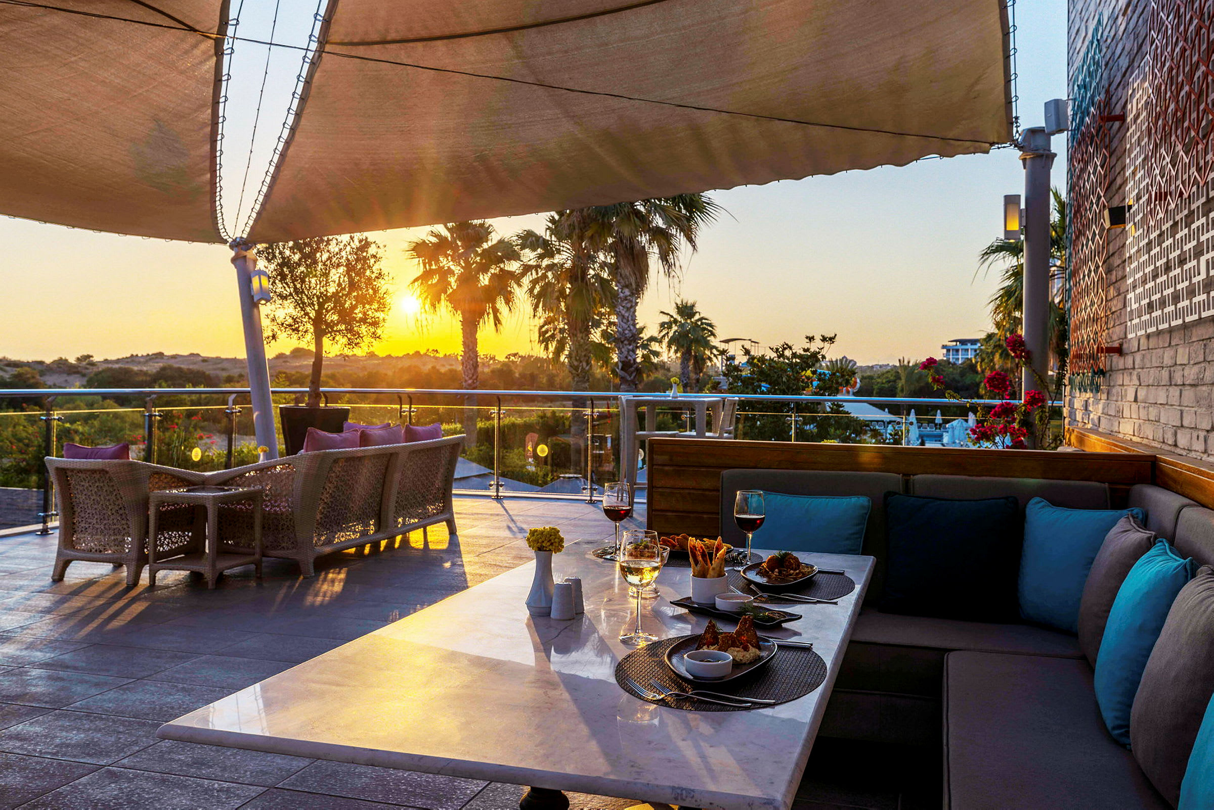 En terrasse med bord og stoler, mat og drikke på et bord, med utsikt over en hage og en solnedgang. (AI-generert tekst)