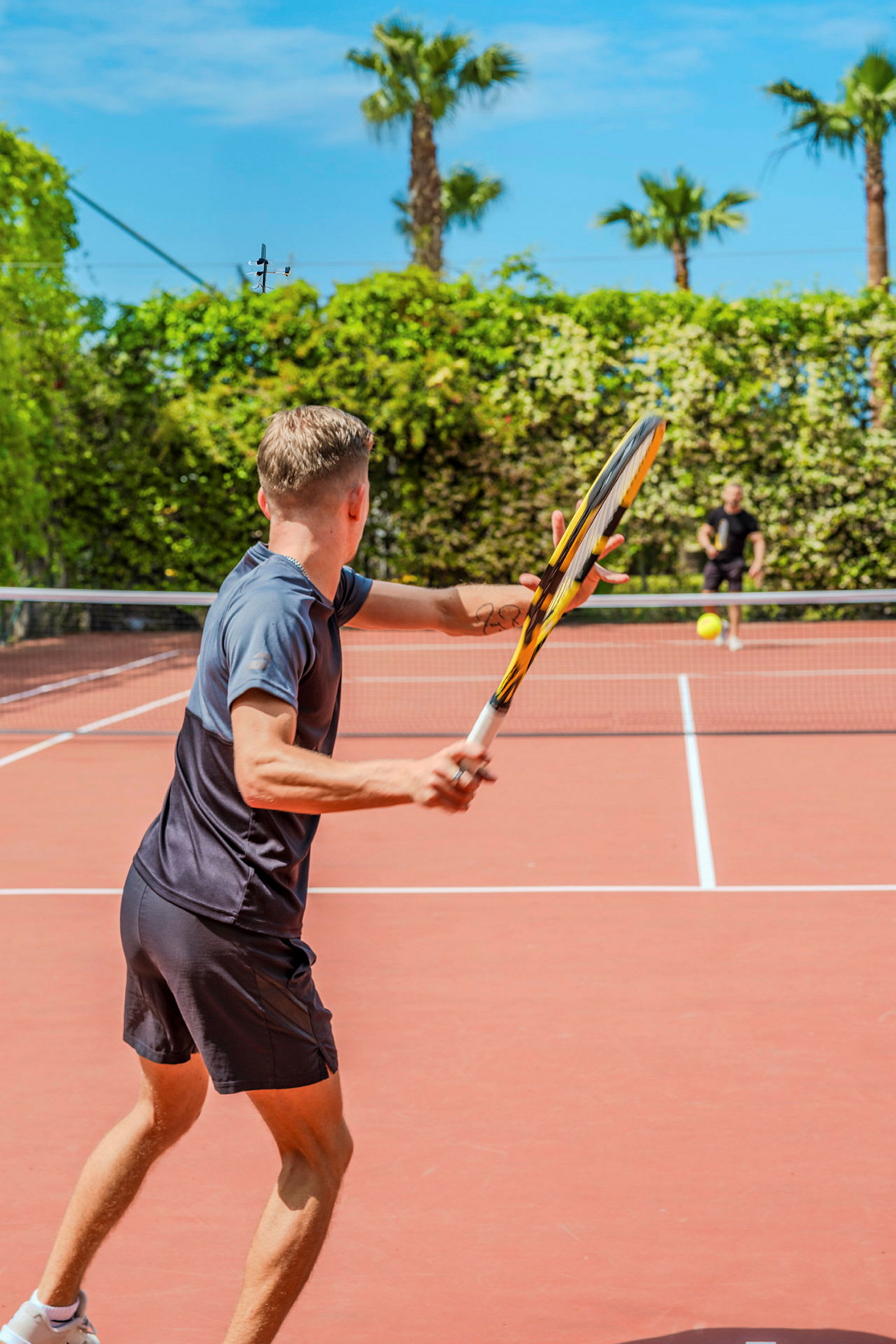 To personer spiller tennis utendørs på en tennisbane omgitt av grøntområde og palmer. (AI-generert tekst)