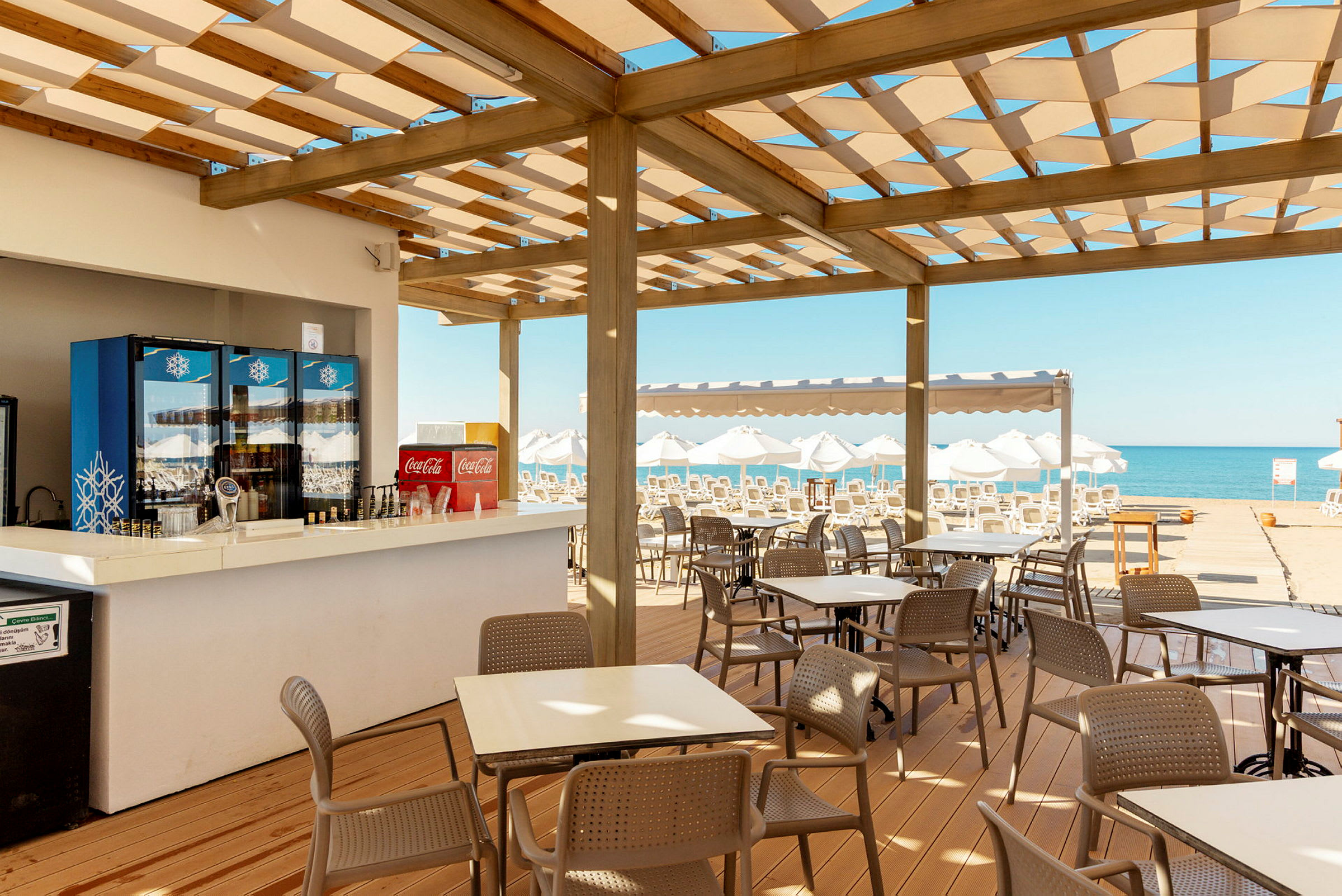 En kafé med uteservering under tretak nær stranden. Bord og stoler er plassert ut, og i bakgrunnen sees solsenger og havet. (AI-generert tekst)