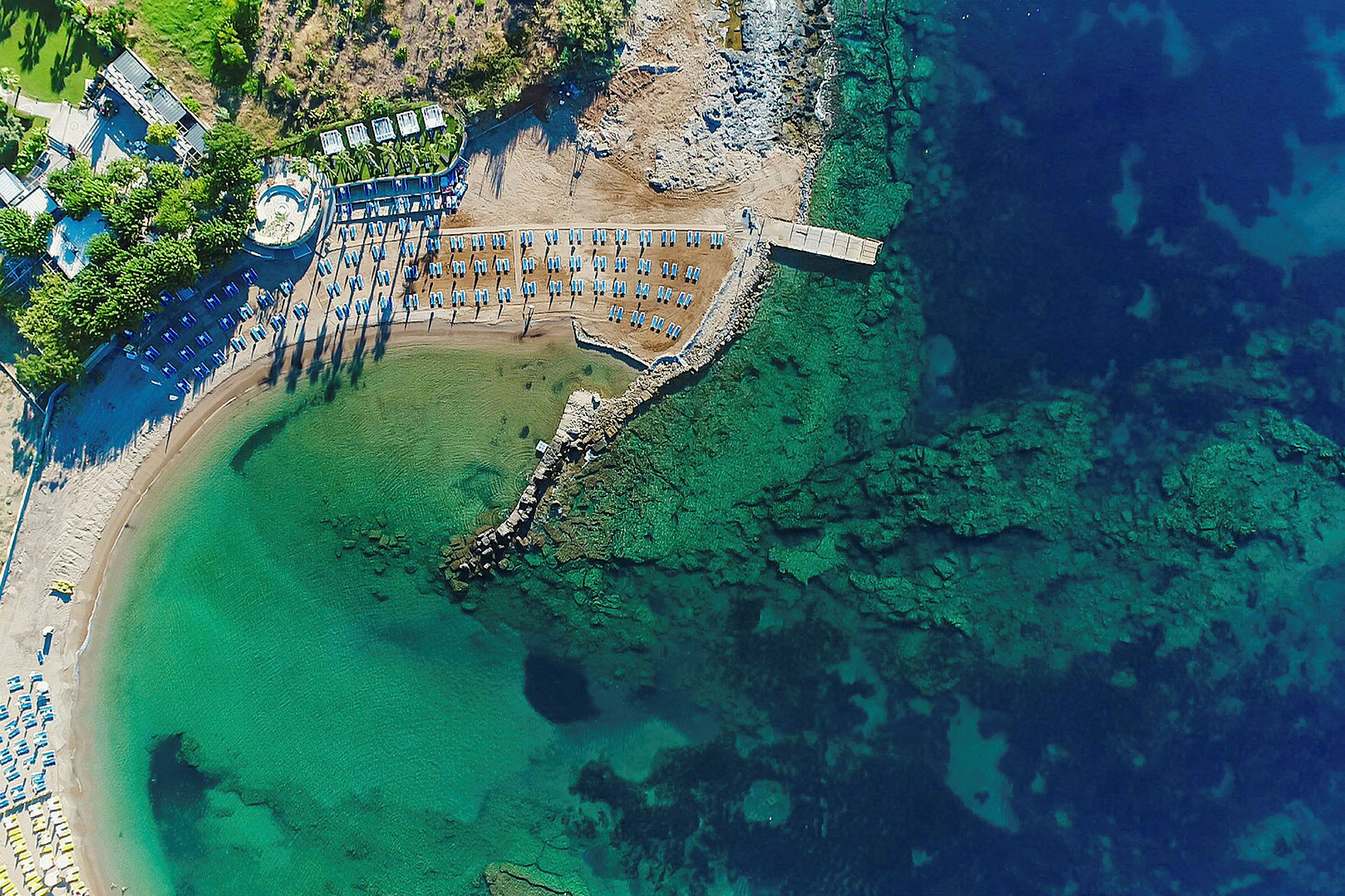 Flybilde av en strand med solsenger og parasoller ved et turkis hav. (AI-generert tekst)