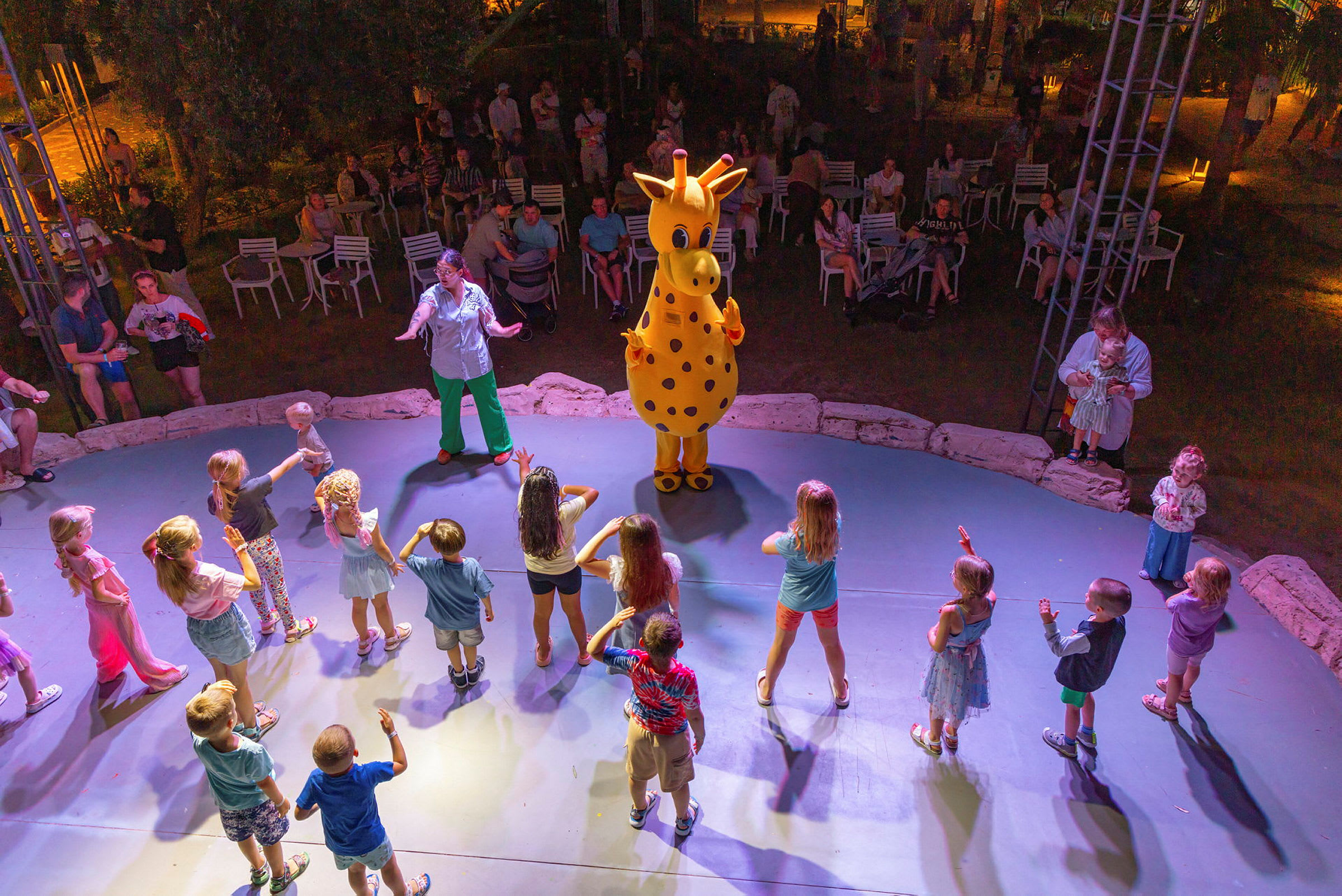 En gruppe barn danser rundt en maskot som forestiller en gul giraff på en scene, mens voksne sitter og ser på i bakgrunnen. (AI-generert tekst)