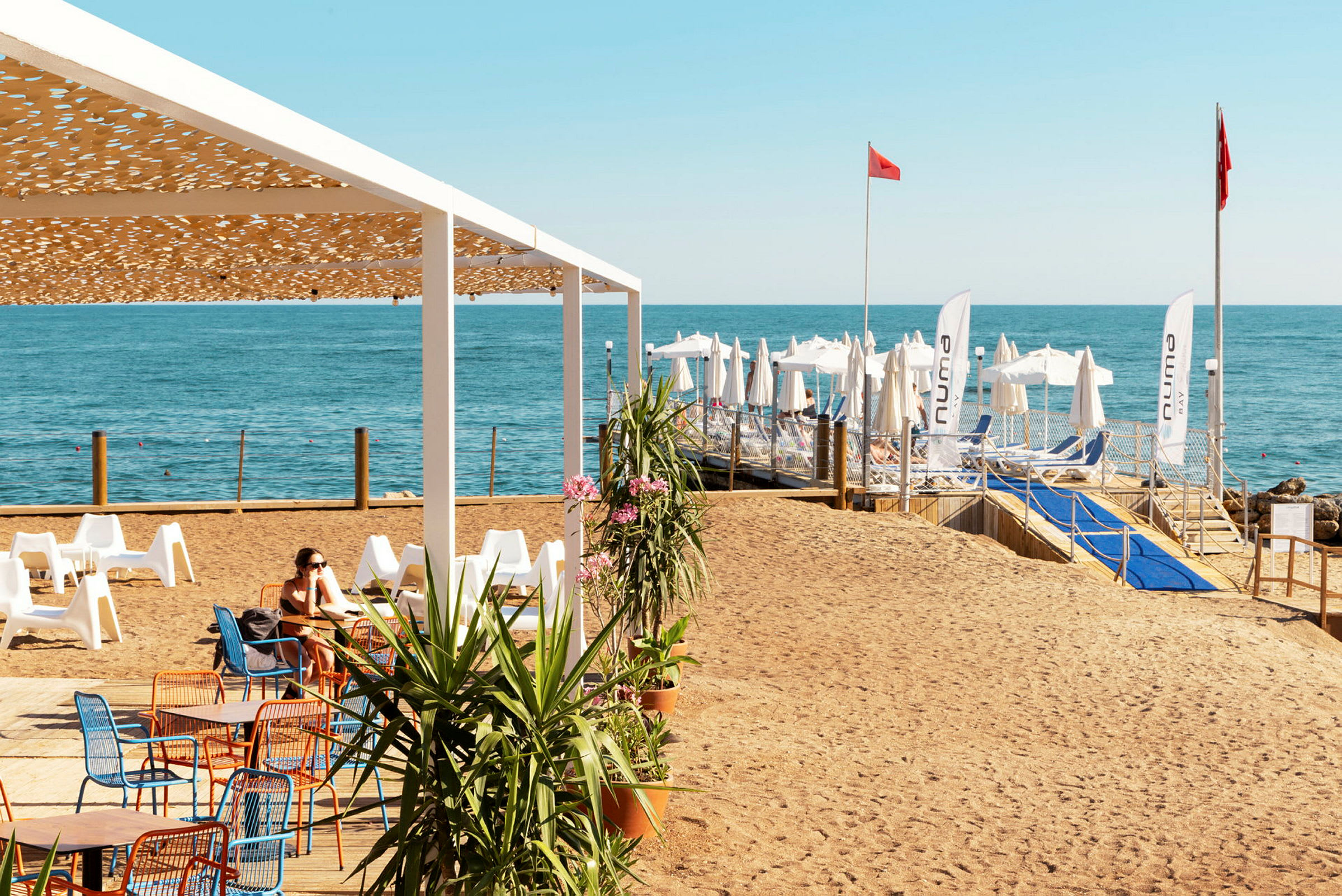 En strand med stoler og bord under en pergola, og i bakgrunnen sees solsenger og parasoller ved havet. (AI-generert tekst)