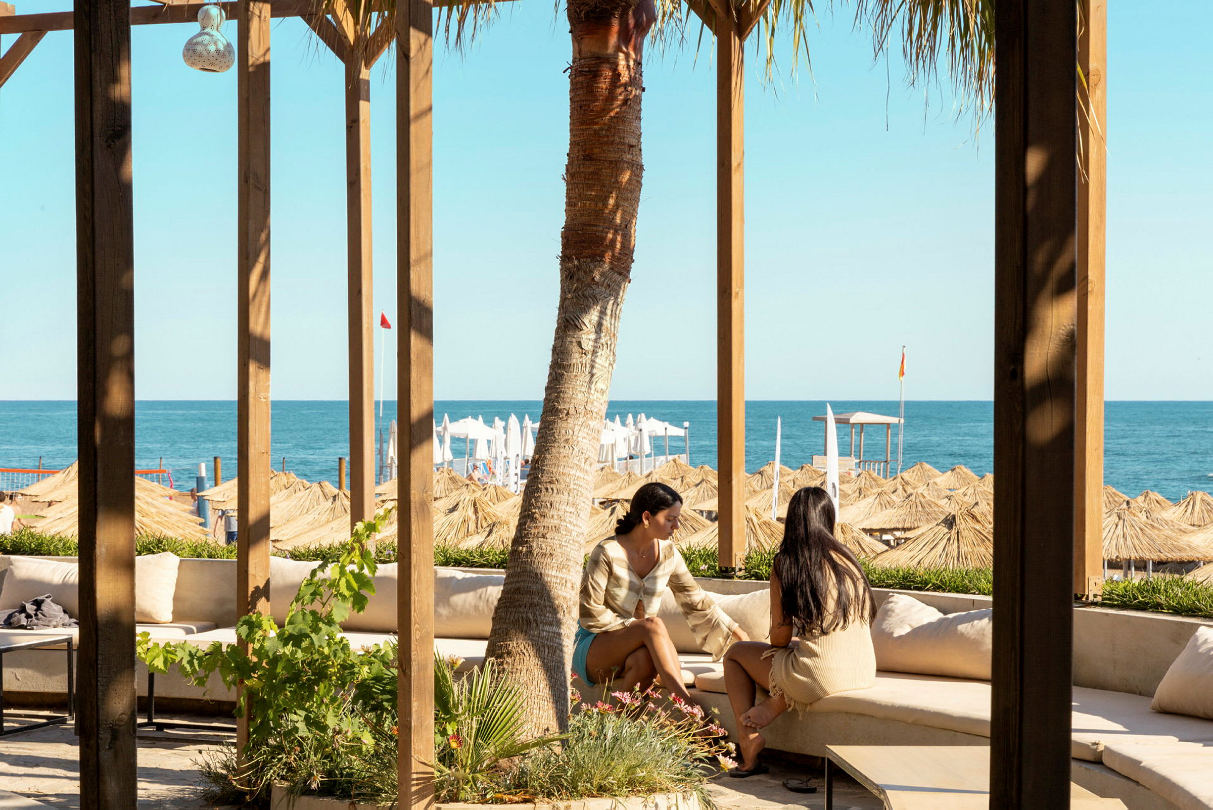 To personer sitter på en utendørssofa under en trepergola og ser utover havet og stranden med parasoller. (AI-generert tekst)