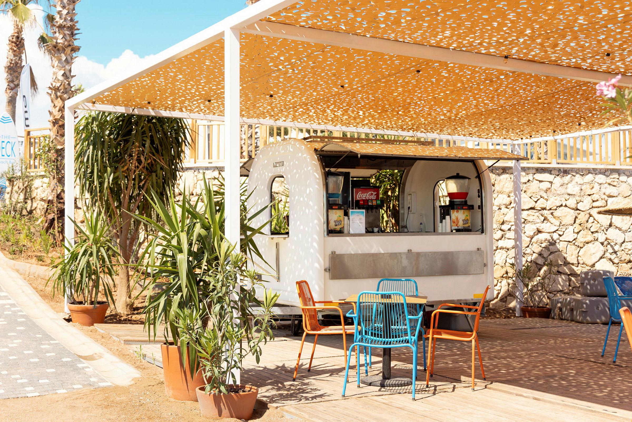 En utendørskafé med en hvit serveringsvogn under et pergolatak dekorert med mønstre. Foran står bord og stoler i forskjellige farger, omgitt av planter i potter. (AI-generert tekst)