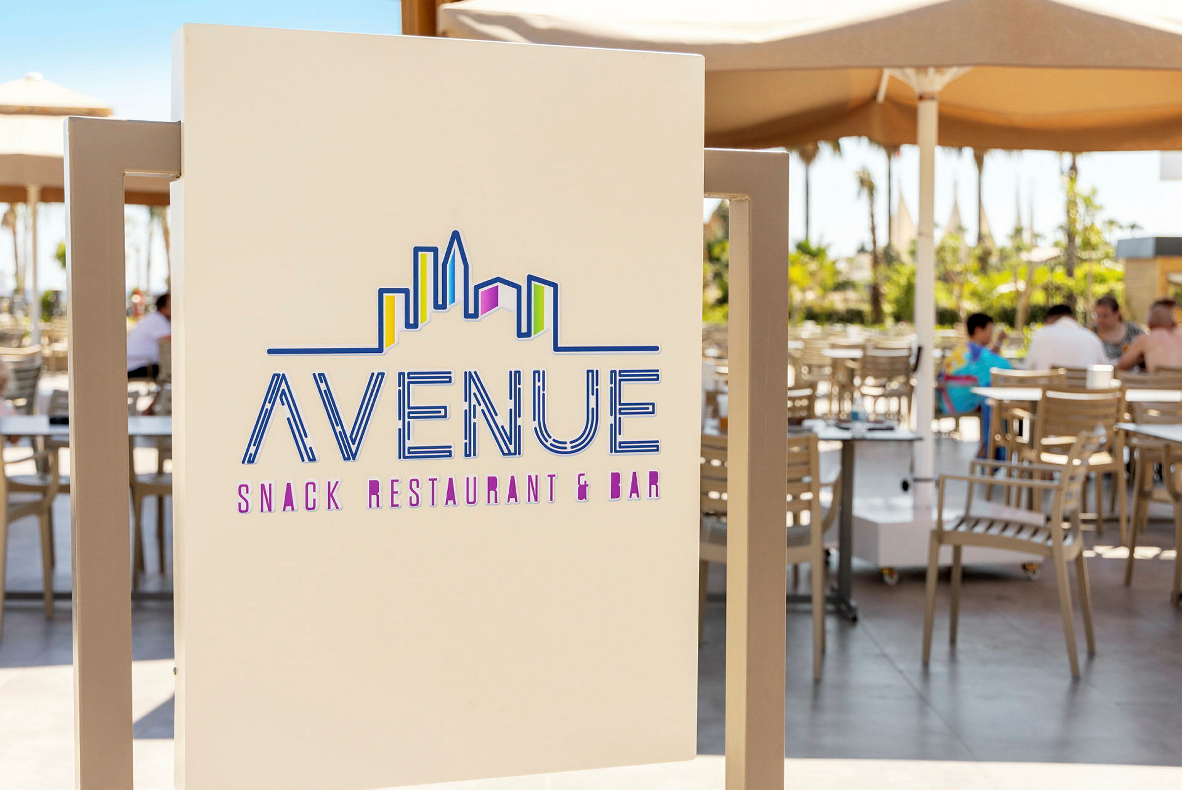 Et skilt som viser teksten "Avenue Snack Restaurant & Bar" med et stilisert skyline-motiv. I bakgrunnen ses en uteservering med bord og stoler under parasoller. (AI-generert tekst)