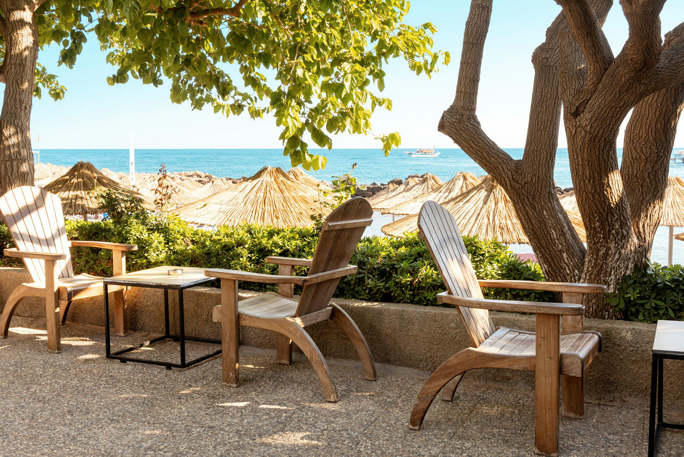 Trestoler og bord står på en terrasse med utsikt over en strand og havet, omgitt av trær og parasoller. (AI-generert tekst)