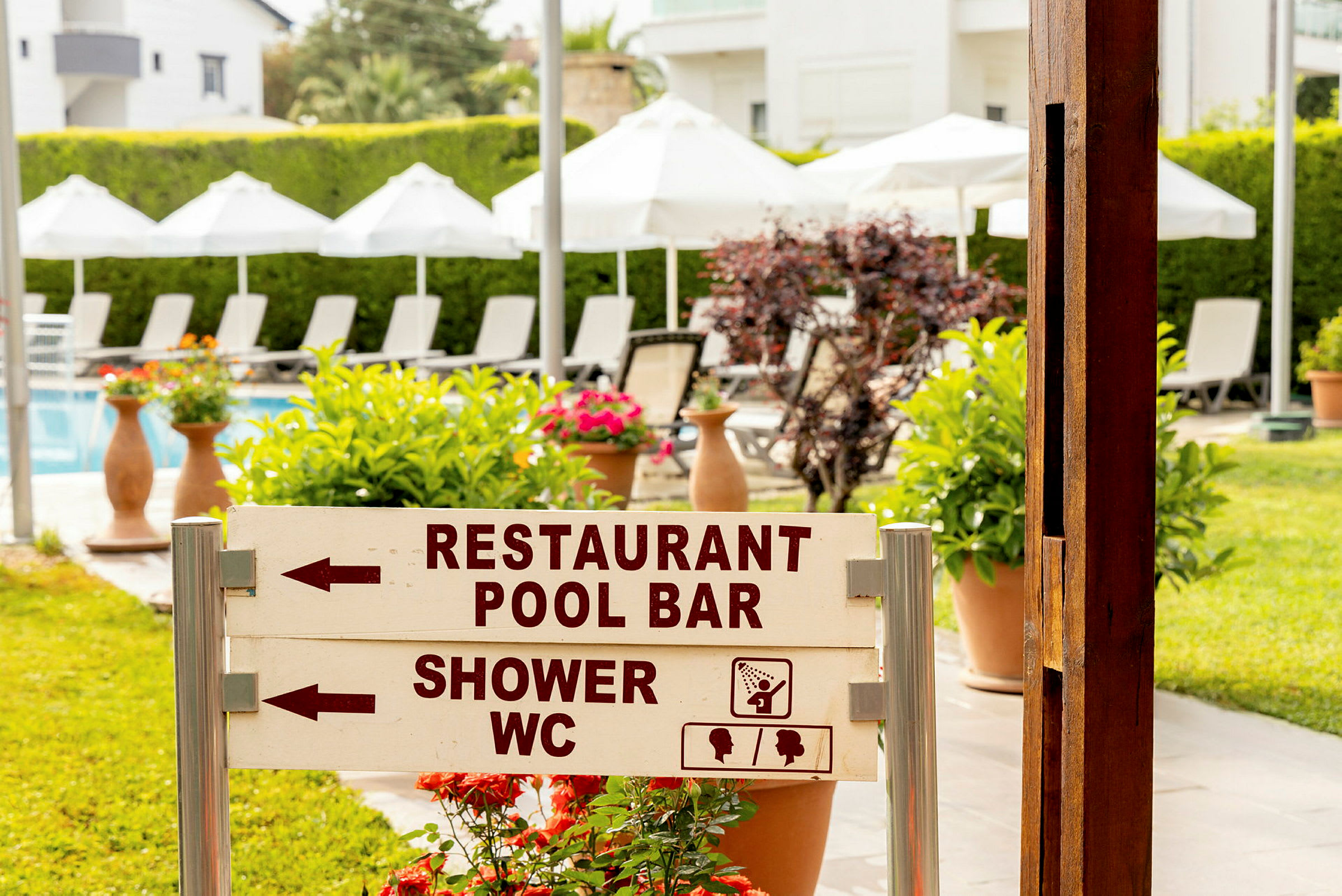 Skilt i en hage med teksten 'Restaurant Pool Bar' og 'Shower WC', solsenger og parasoller vises i bakgrunnen. (AI-generert tekst)