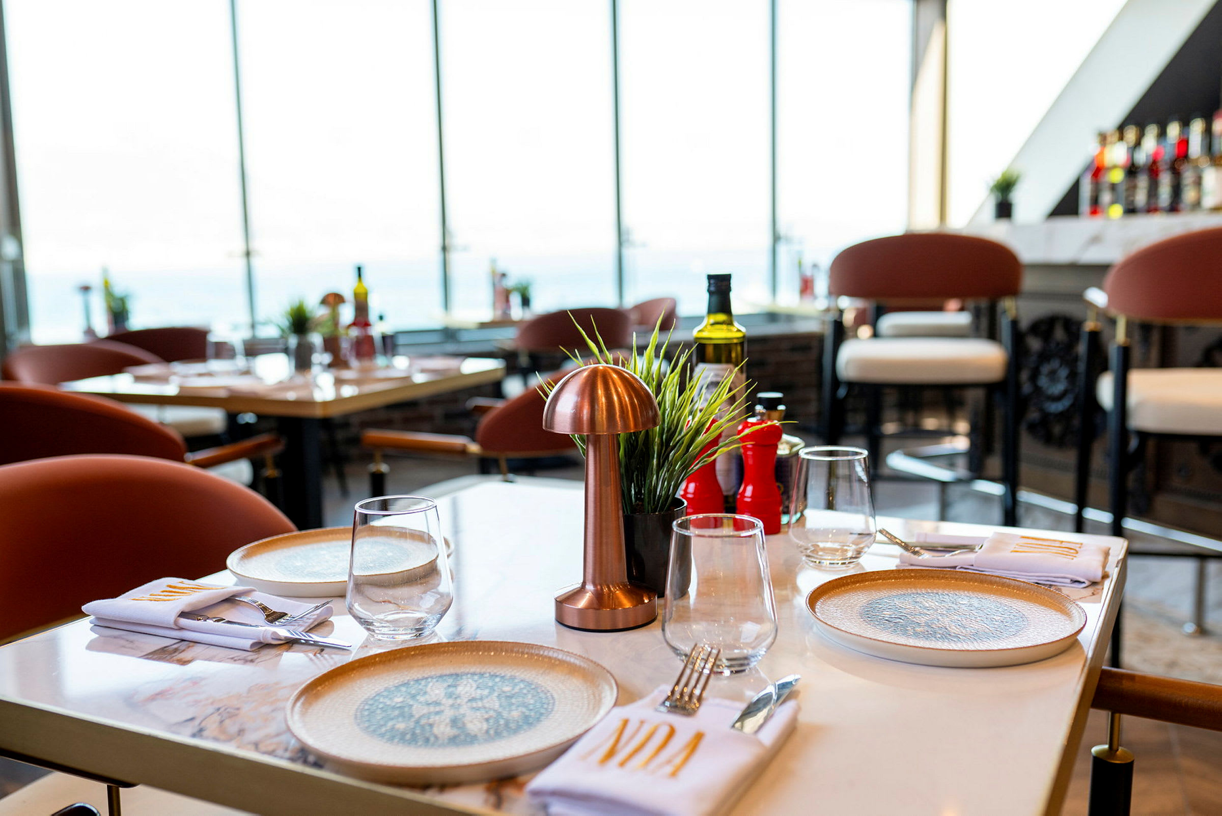 Bord på en restaurant med tallerkener, glass og bestikk, utsikt over store vinduer. (AI-generert tekst)