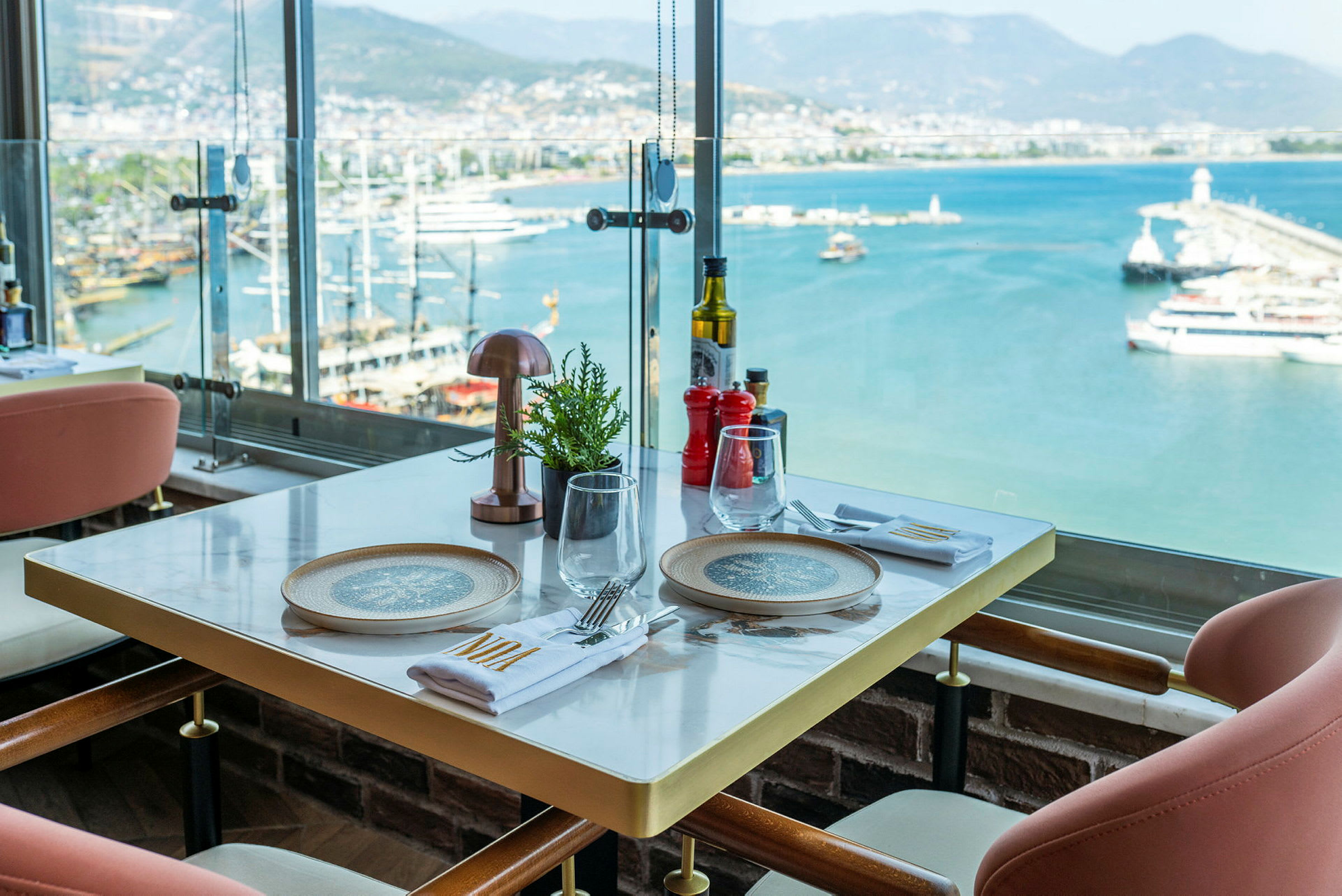 Utsikt fra et restaurantbord med dekking, med en havn og havet i bakgrunnen. (AI-generert tekst)