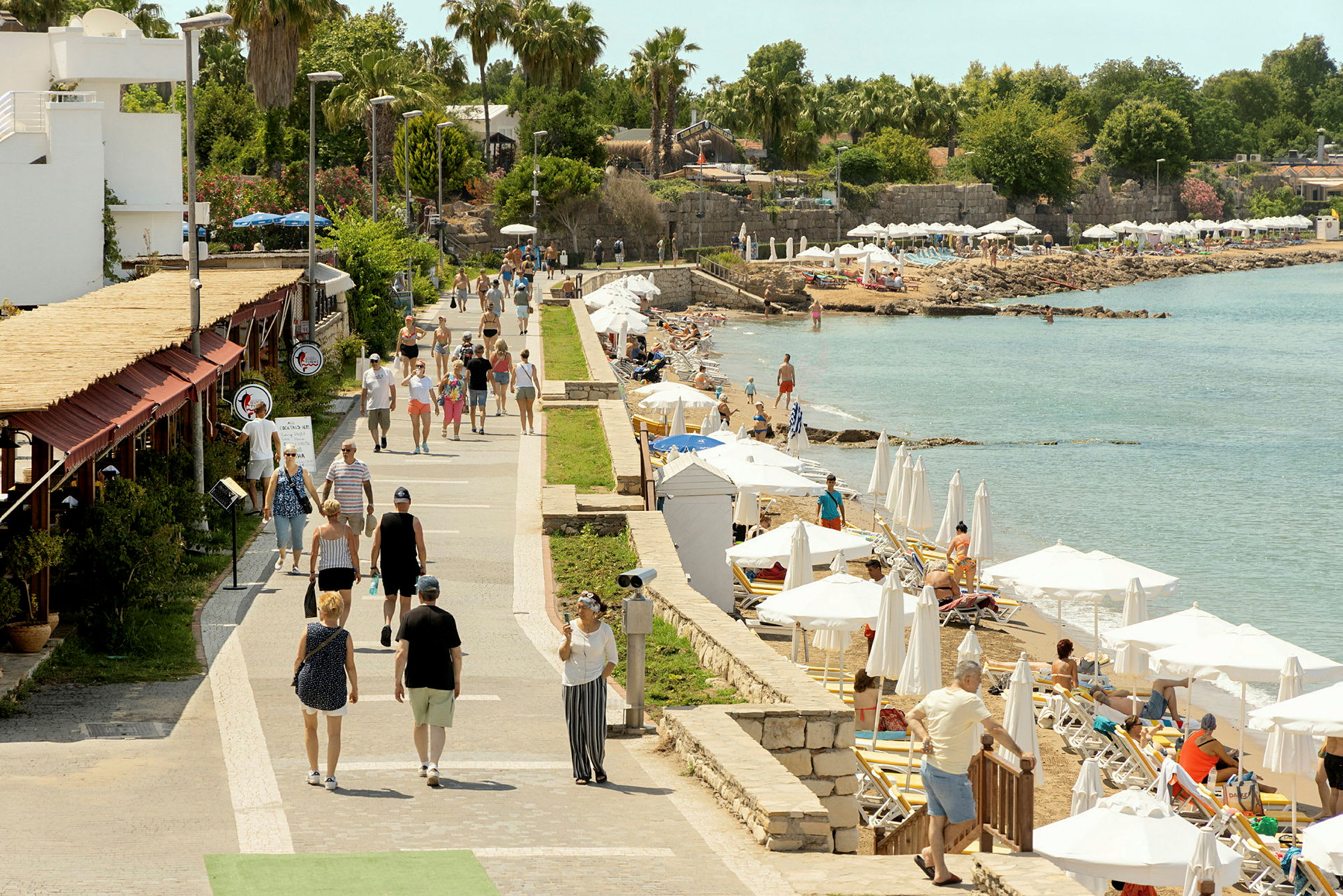 En gangsti ved havet med folk som spaserer og personer som slapper av på solsenger under parasoller. (AI-generert tekst)