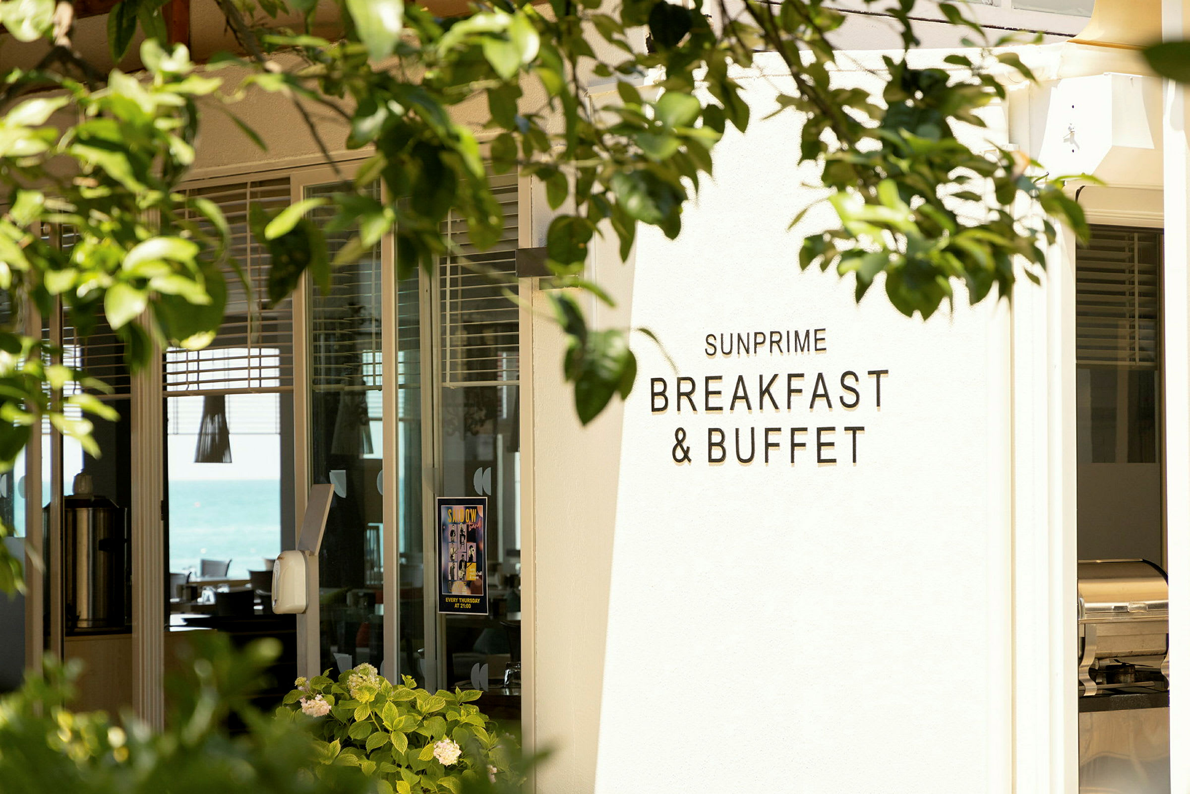 Skilt med teksten 'Sunprime Breakfast & Buffet' ved inngangen til en restaurant, omgitt av grøntområder. (AI-generert tekst)