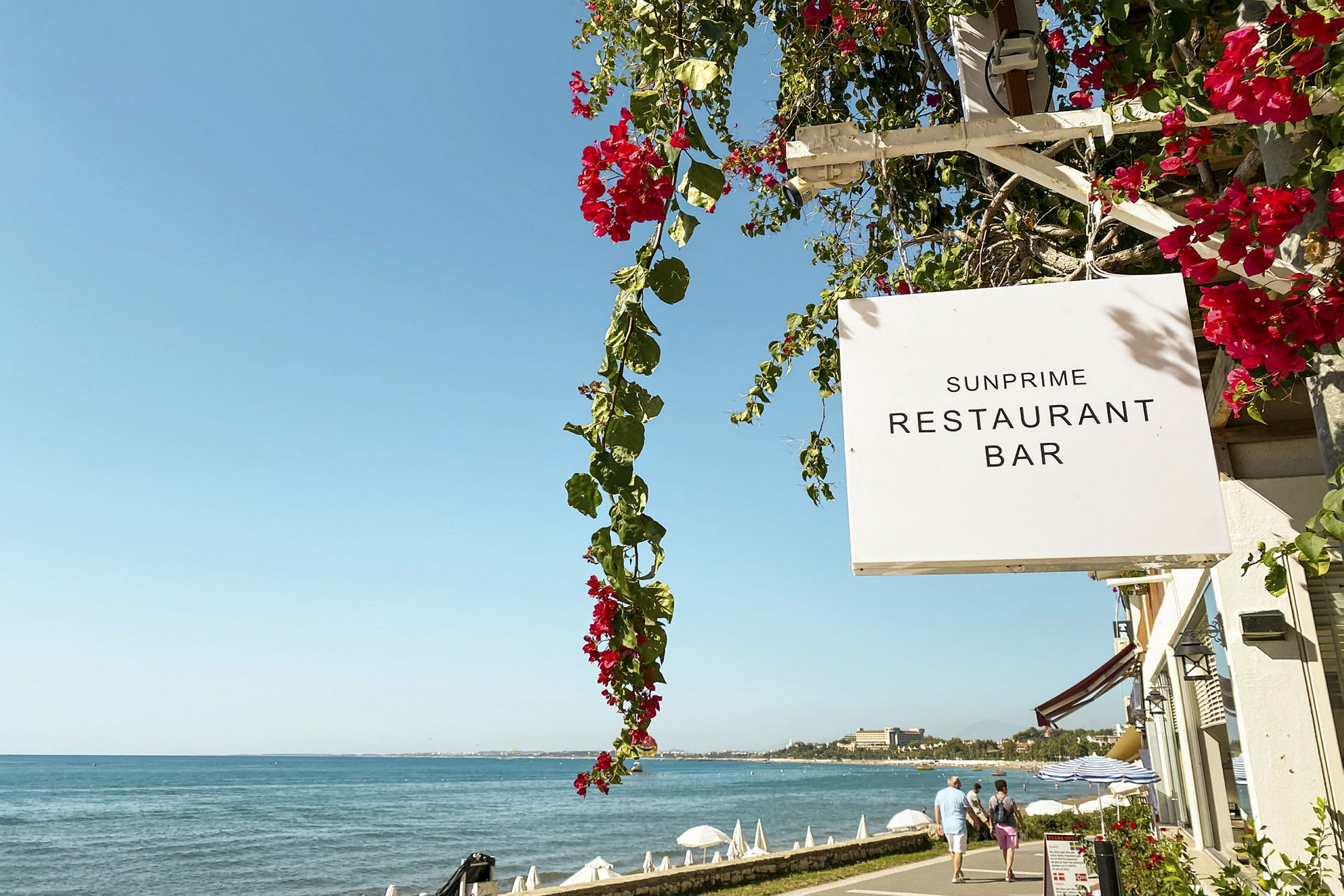 Skilt for Sunprime Restaurant Bar ved en promenade nær sjøen. (AI-generert tekst)