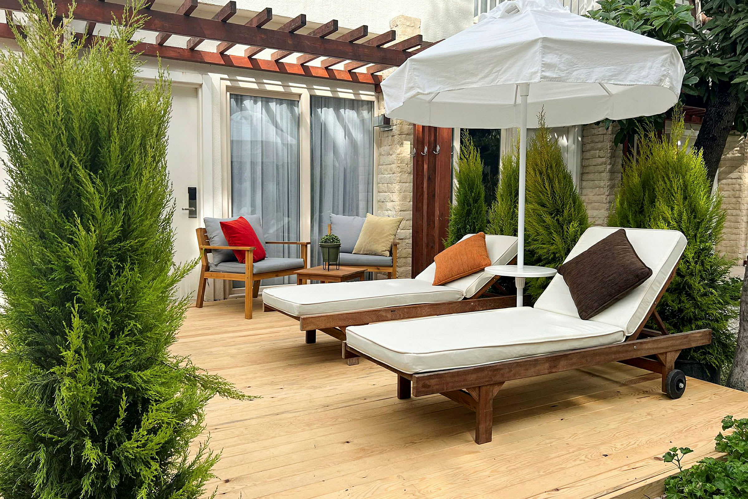 To solsenger med hvite puter og puter står på en treterrasse under en parasoll, omgitt av grønne planter. (AI-generert tekst)