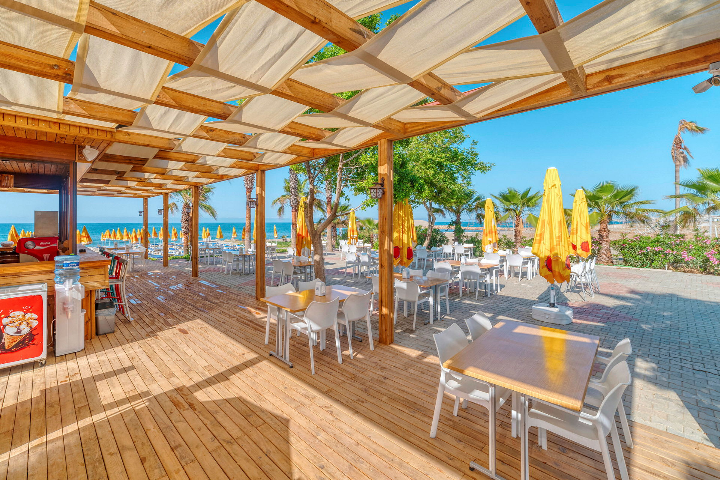 Utendørs spisested med bord og stoler under en pergola ved stranden. (AI-generert tekst)